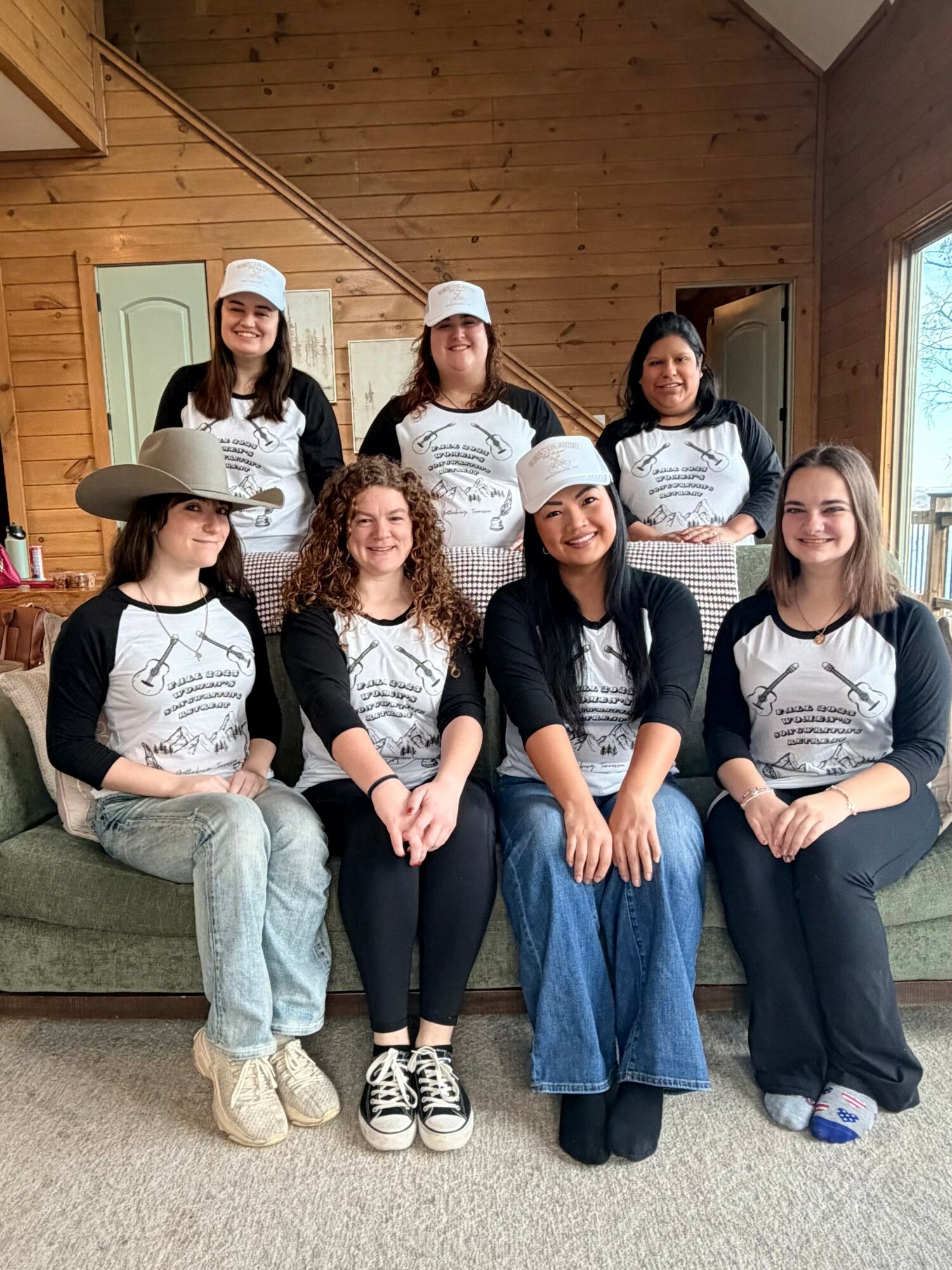 Seven women sitting and standing on a couch in a wooden room, wearing matching shirts and hats, smiling.