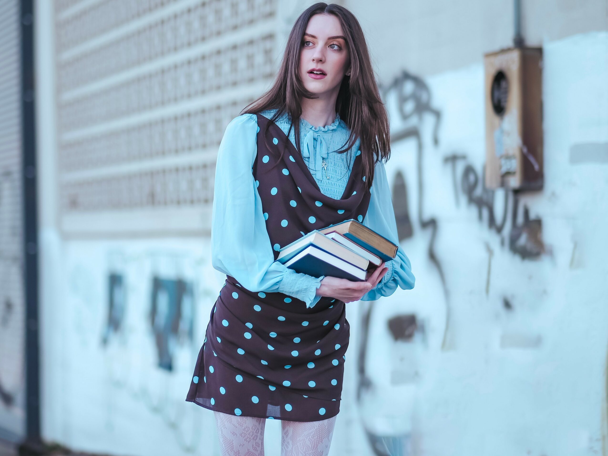 Young woman with long brown hair holding books, standing outdoors near graffiti-covered wall, wearing a blue blouse and polka dot skirt.