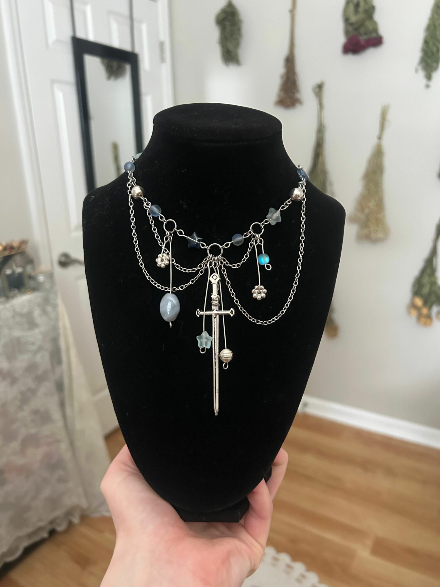 Black jewelry display bust with layered silver necklaces featuring charms and pendants, in a room with wall decorations and wooden floor.