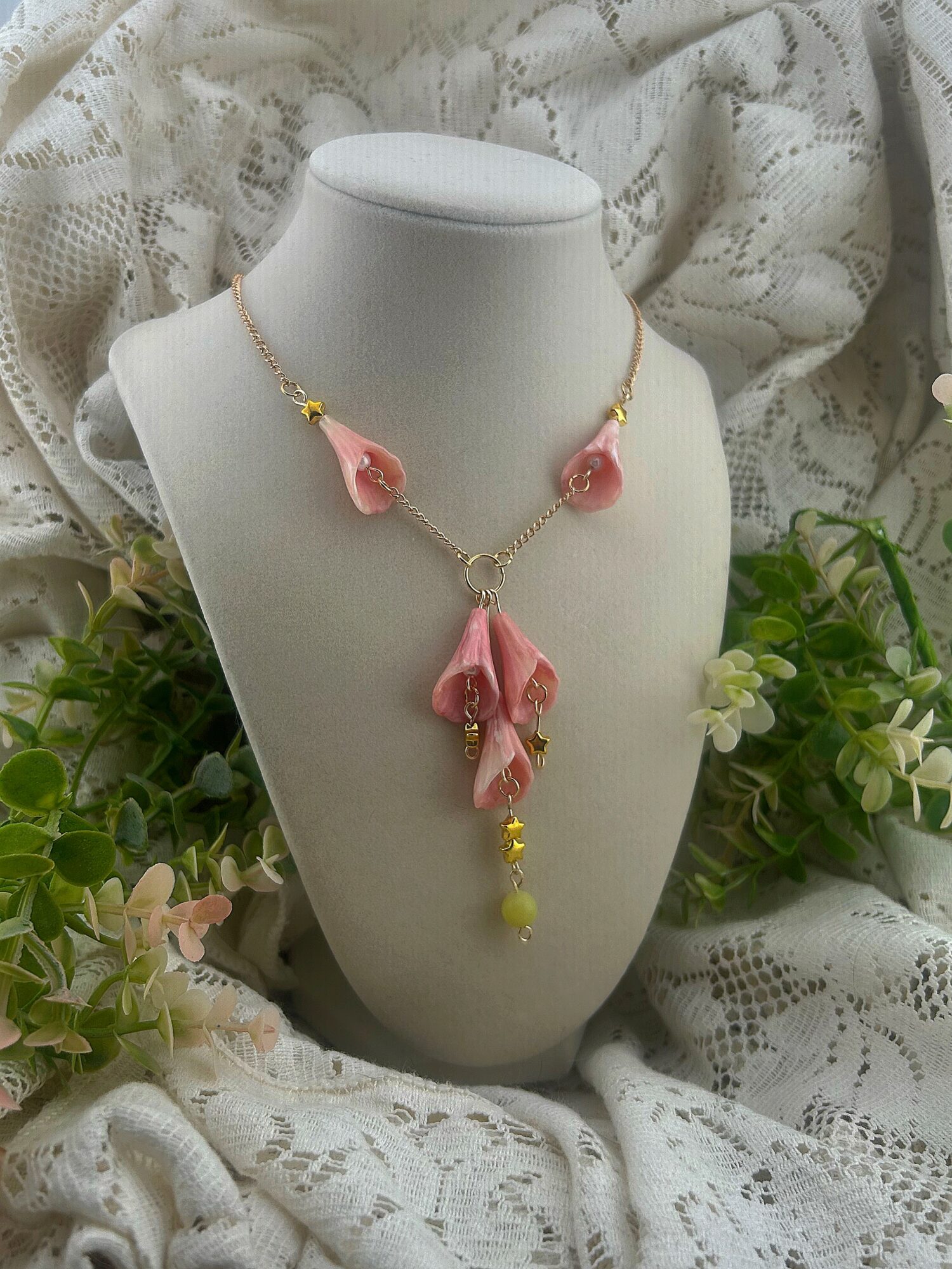 Pink flower-shaped necklace with yellow beads displayed on a white bust, surrounded by lace fabric and green plants.
