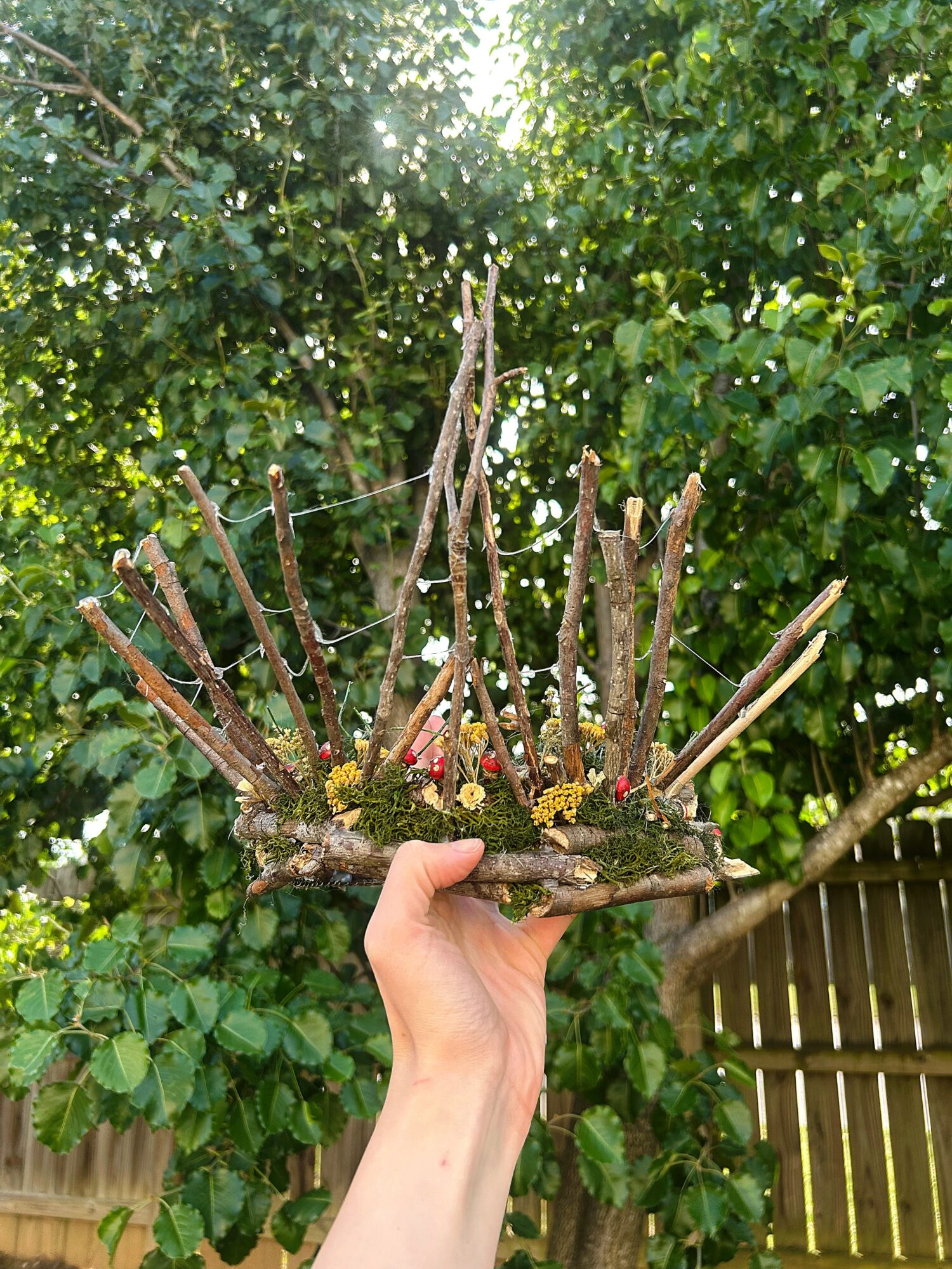 Hand holding a small wooden structure with sticks and flowers, outdoors with green foliage and a wooden fence in background.