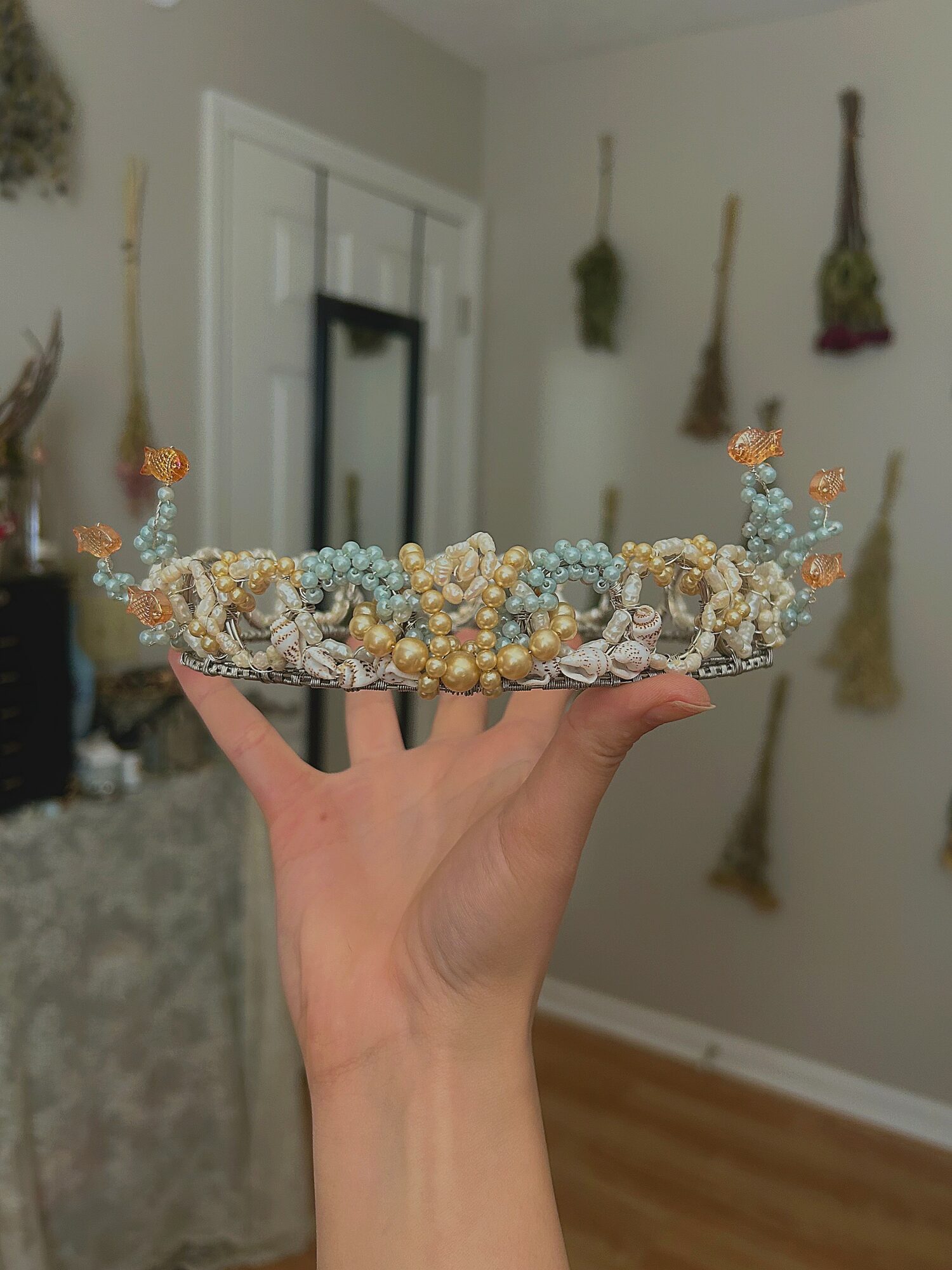 Hand holding a decorative tray with beaded jewelry, in a room with hanging dried herbs or flowers on the wall.