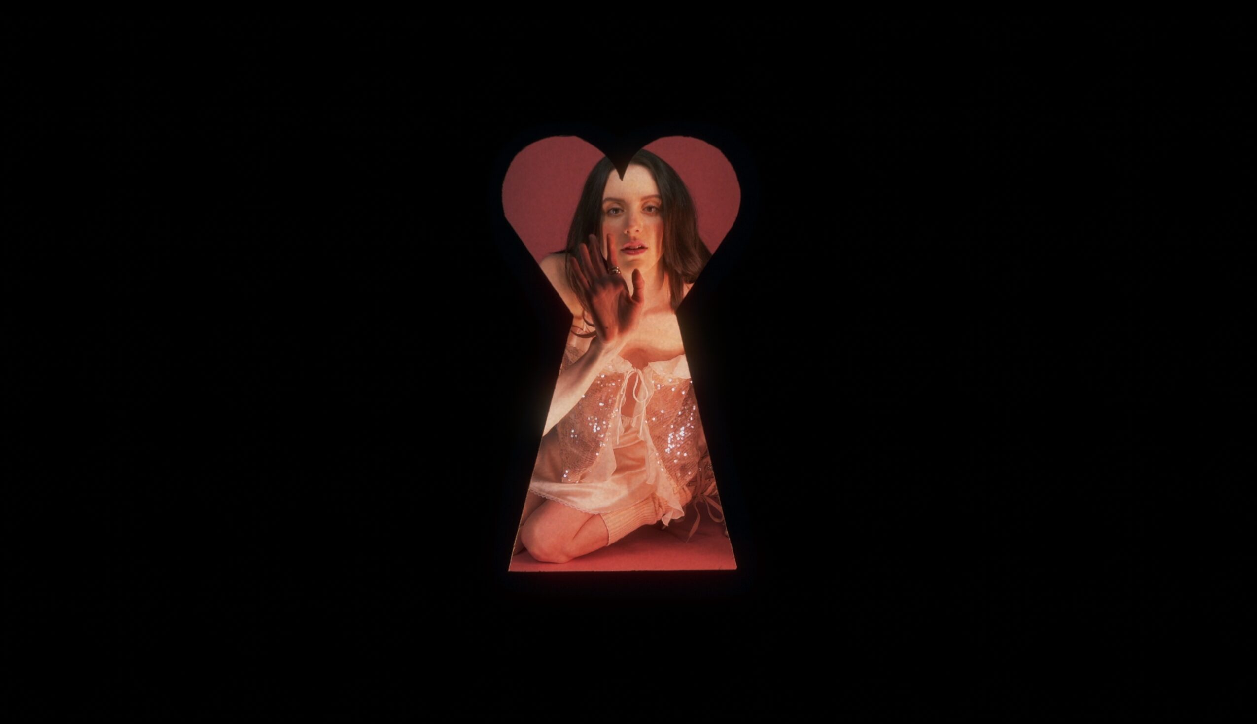 A woman with long dark hair and makeup, looking through a keyhole-shaped opening, with a pink background.