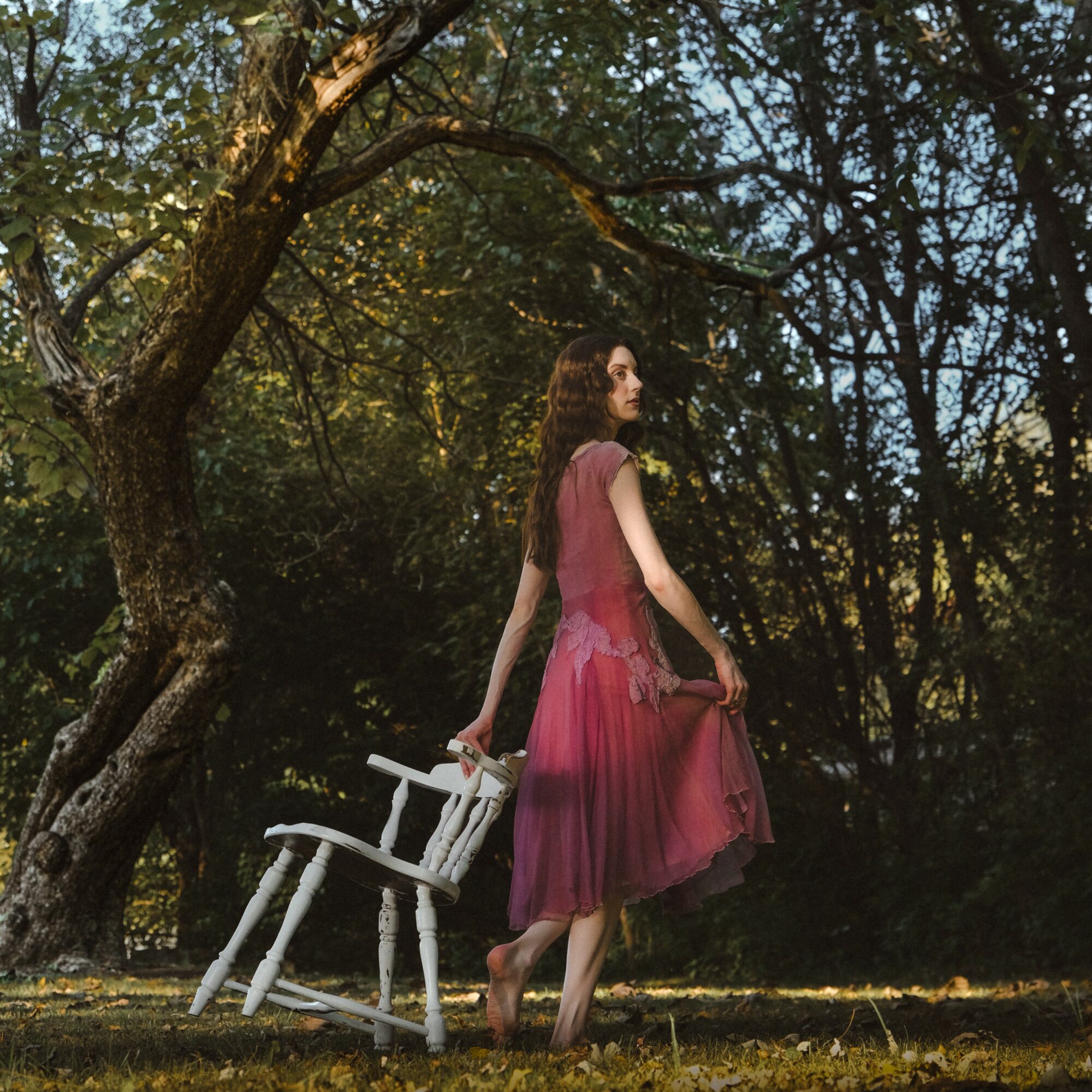 Woman in a pink dress standing outdoors near a white chair and a large tree, surrounded by trees and sunlight.