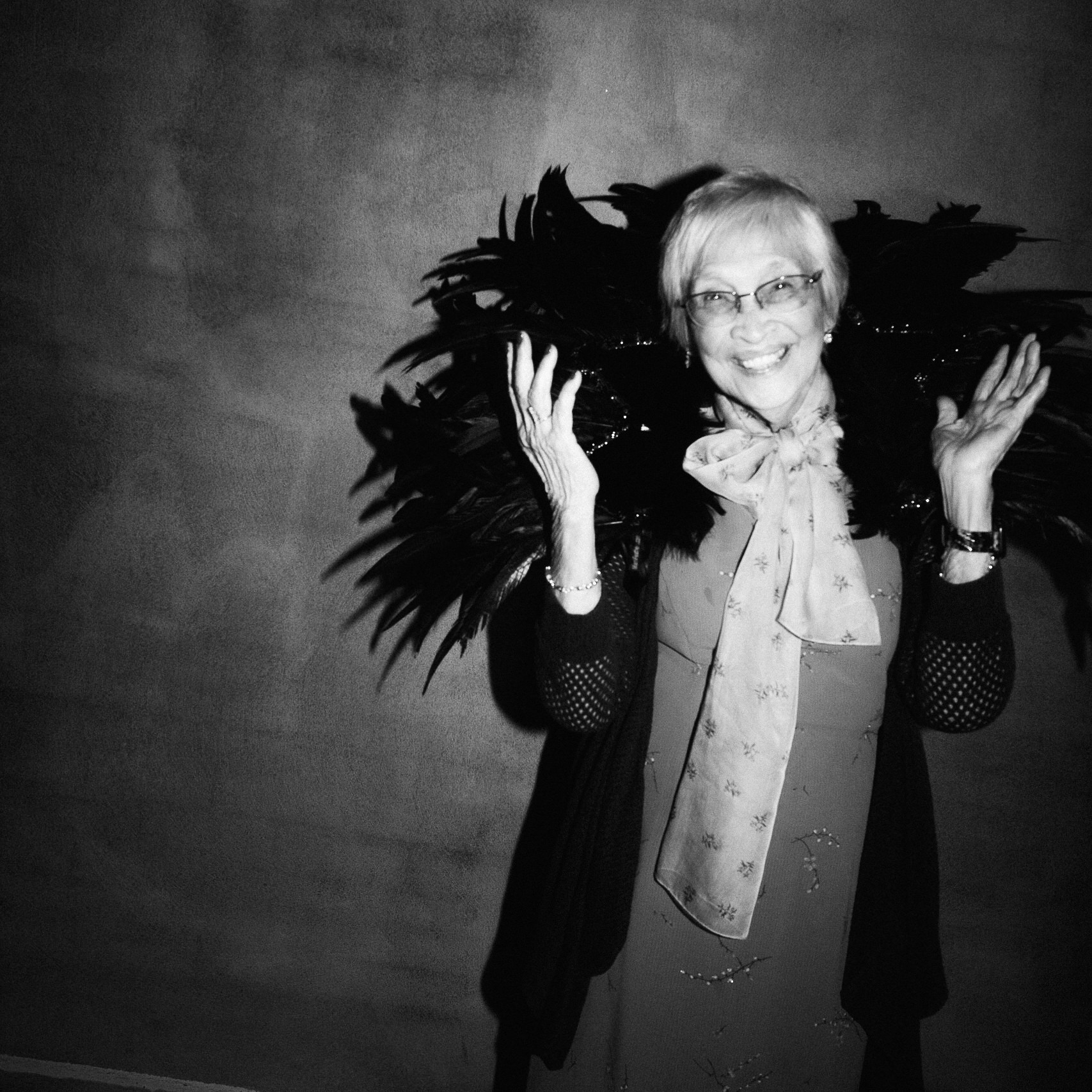 Older woman with glasses smiling, holding a large feathered accessory, standing against a plain background.
