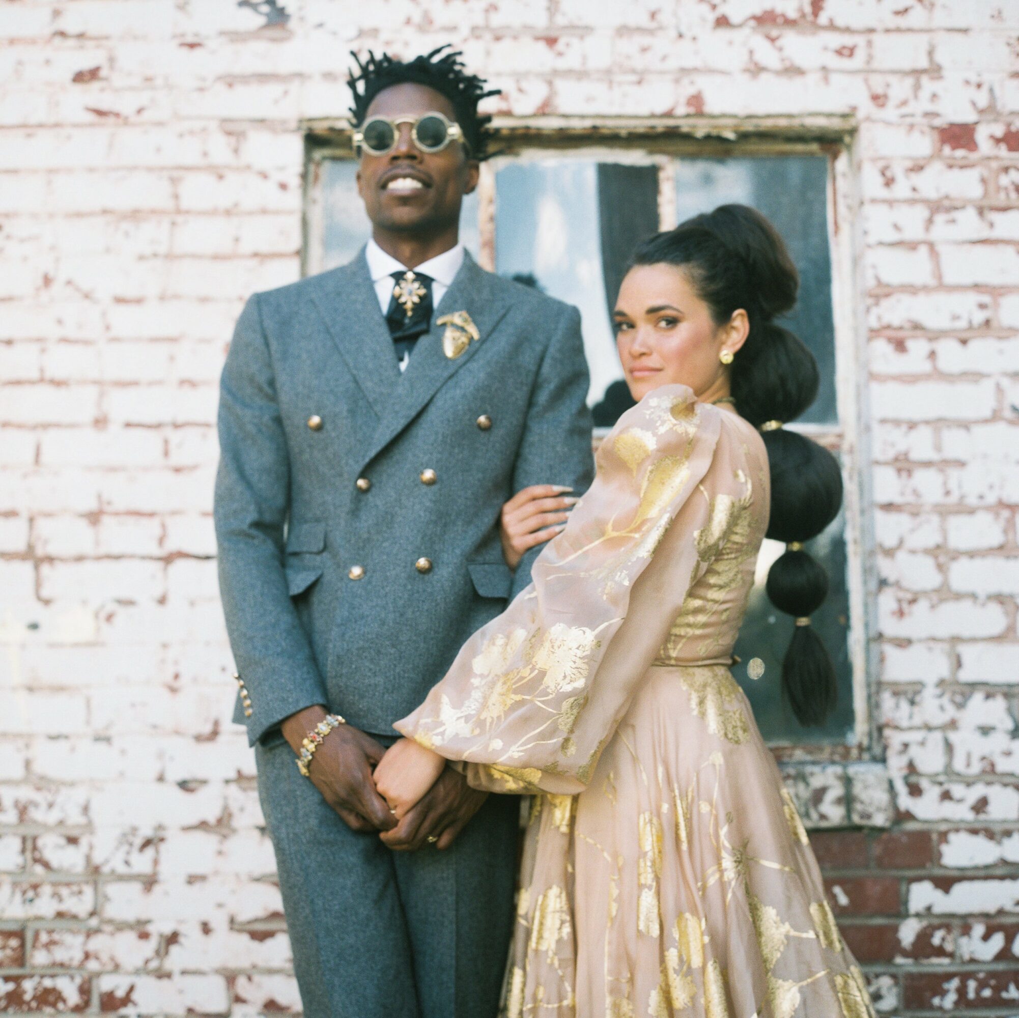 A man and woman stand in front of a brick wall with a window, dressed in vintage-style clothing.