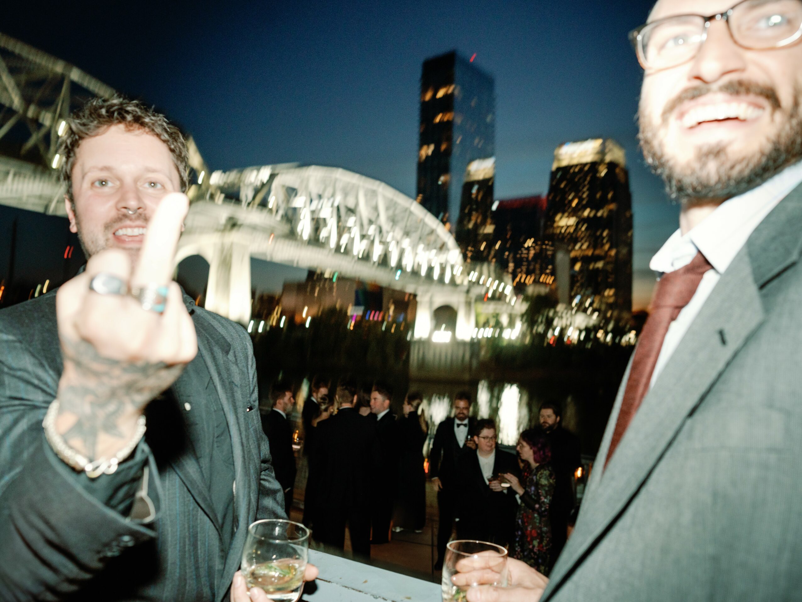 Two men in suits at an outdoor event with a city skyline and illuminated bridge in the background.