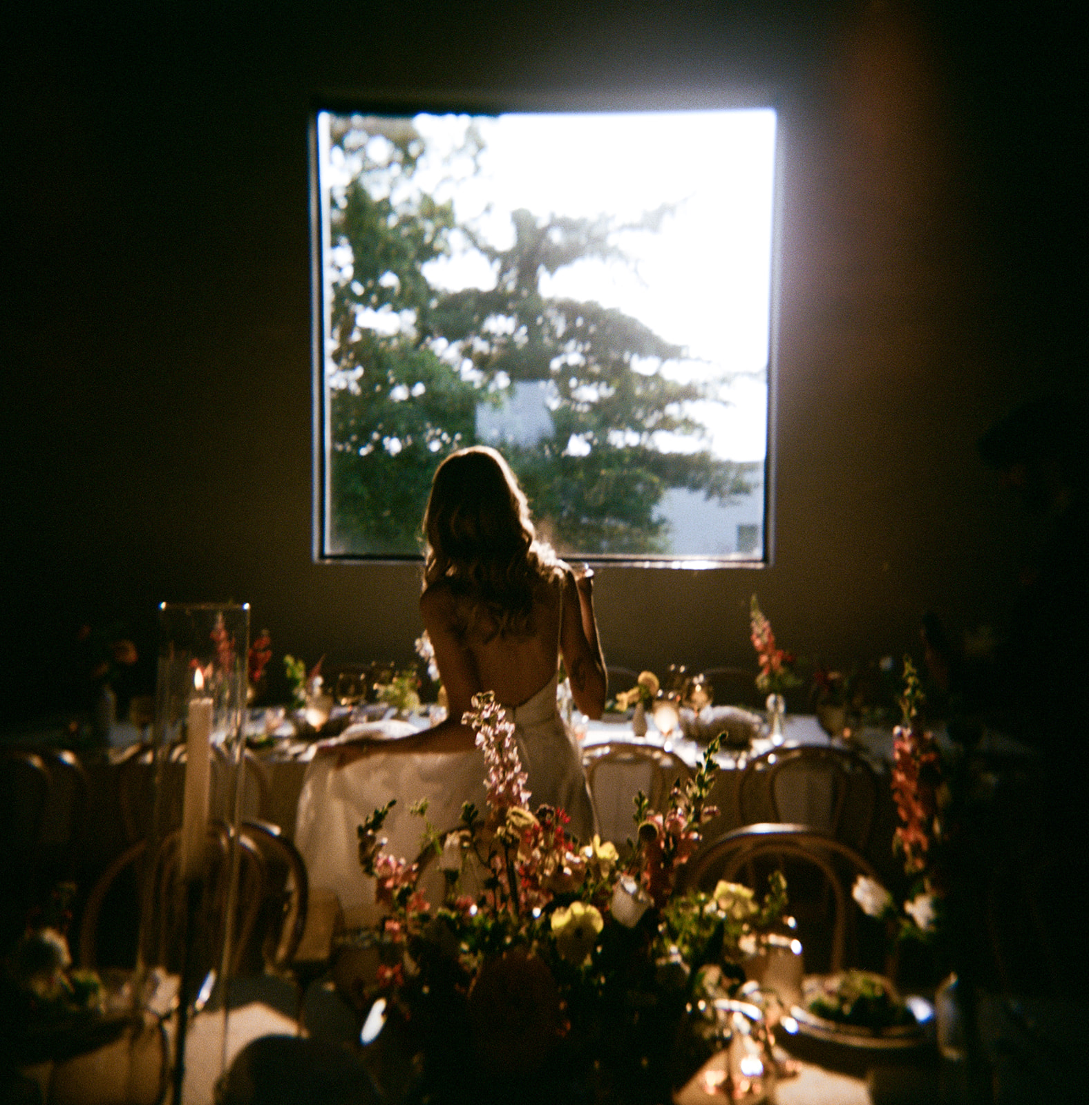 Person with long hair sitting at a table with floral arrangements, looking out a window with trees outside.