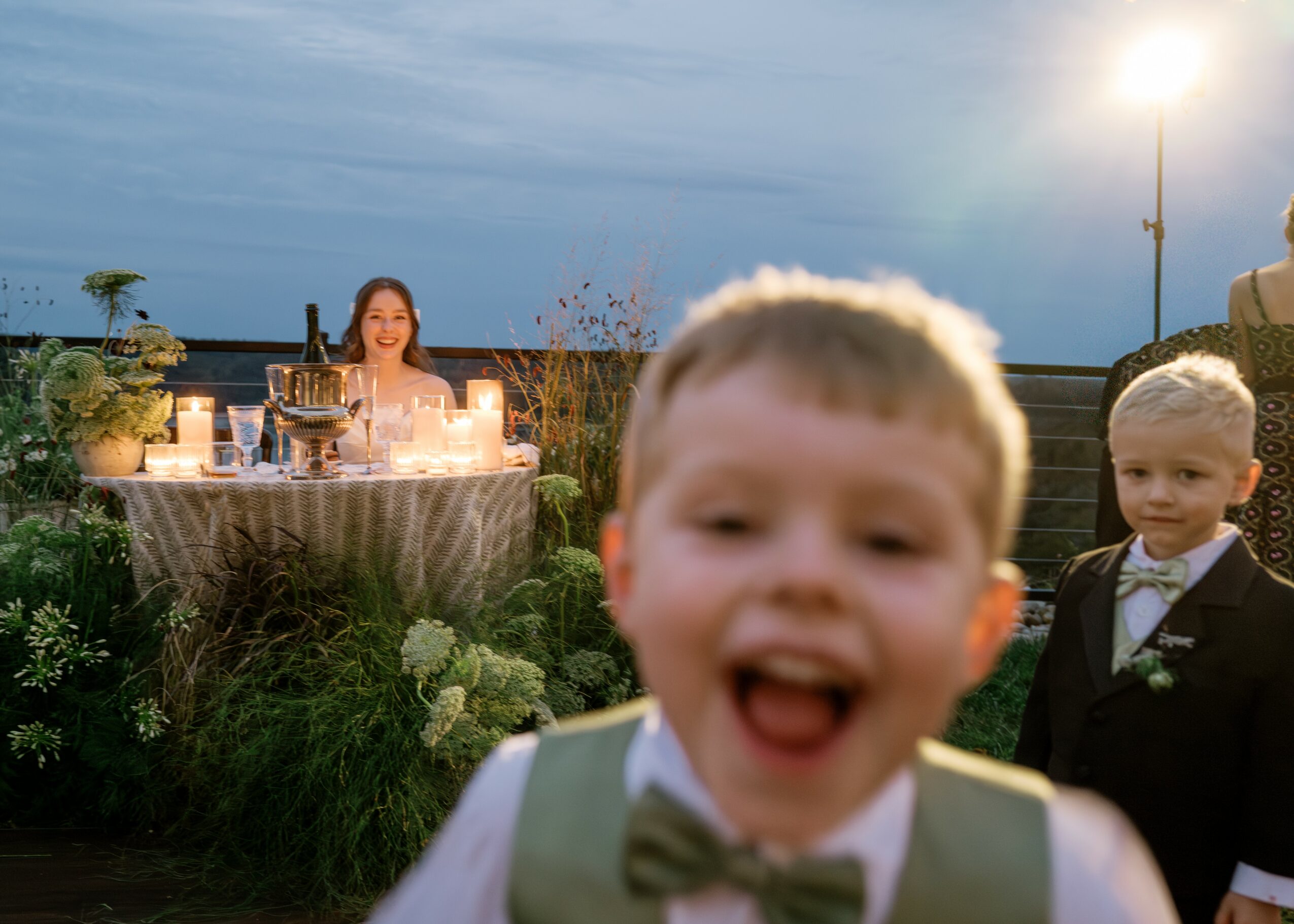 Two children in formal attire smiling outdoors with a woman at a table in the background.