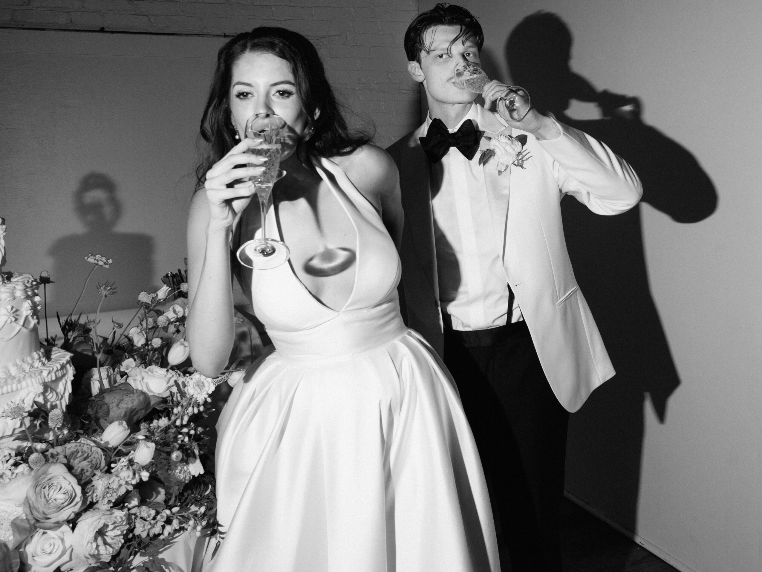 Woman in a dress and man in a tuxedo drinking from glasses at an event with floral arrangements.