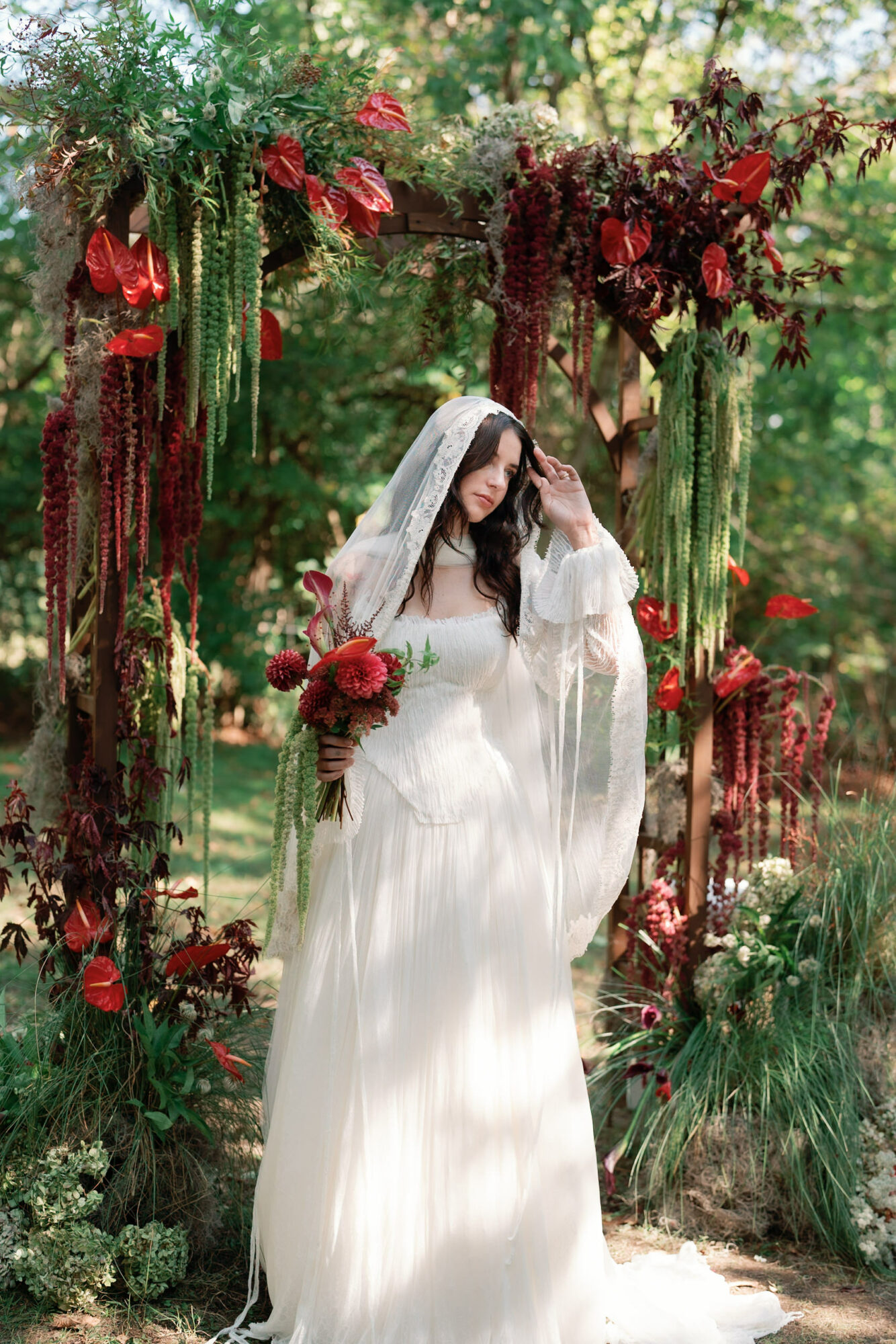 Woman in white dress holding bouquet, standing outdoors with floral arch and greenery.