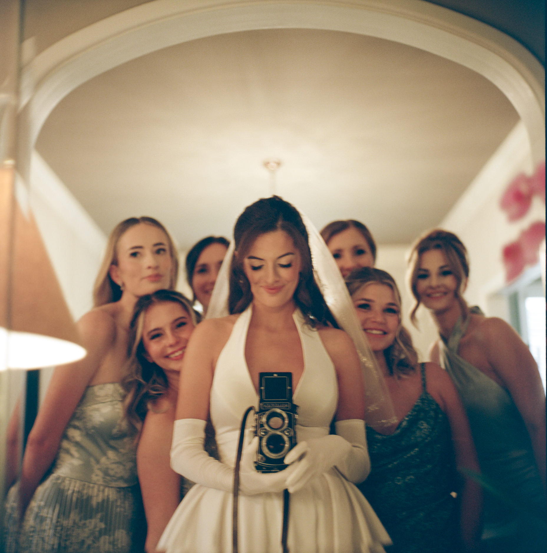 Group of women in elegant dresses, with one woman in the center holding a vintage camera, smiling.