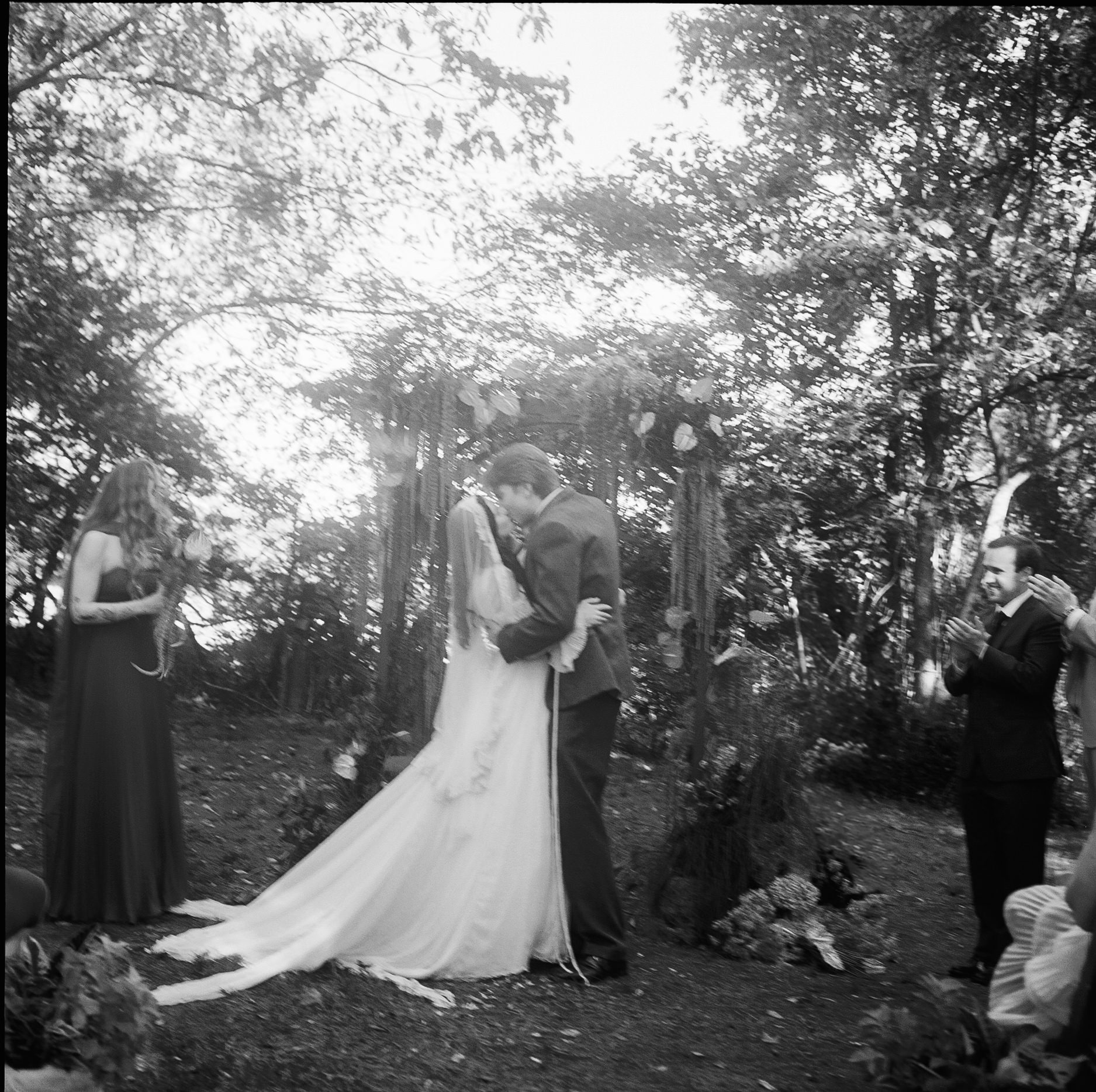 Couple kissing outdoors, surrounded by trees, with two women on either side, one holding a book, in a wedding scene.
