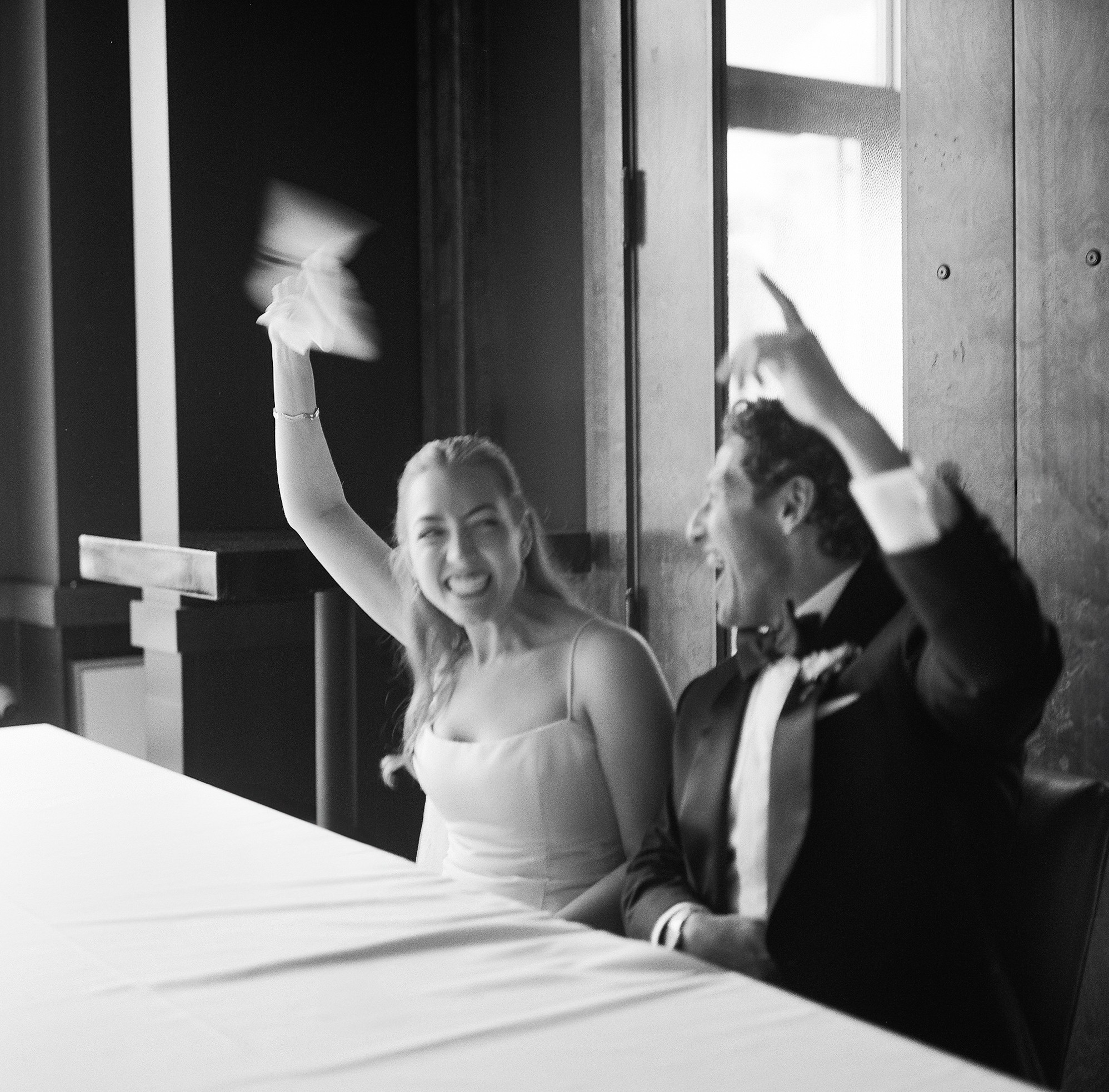 Woman and man in formal attire sitting at a table, woman waving her hand, smiling, in a room with large windows.