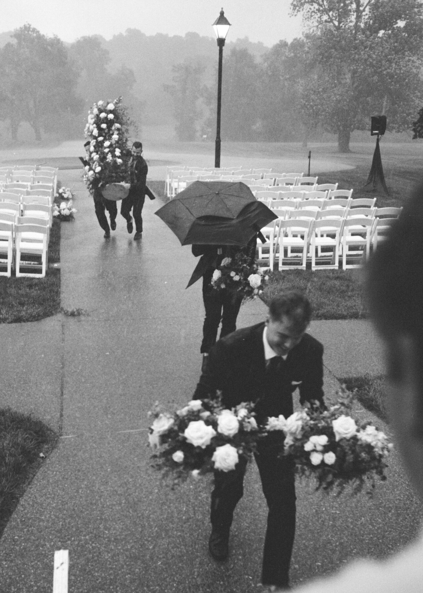 Person carrying a large floral arrangement walking on a wet path, with a person holding an umbrella in the background, outdoor setting.