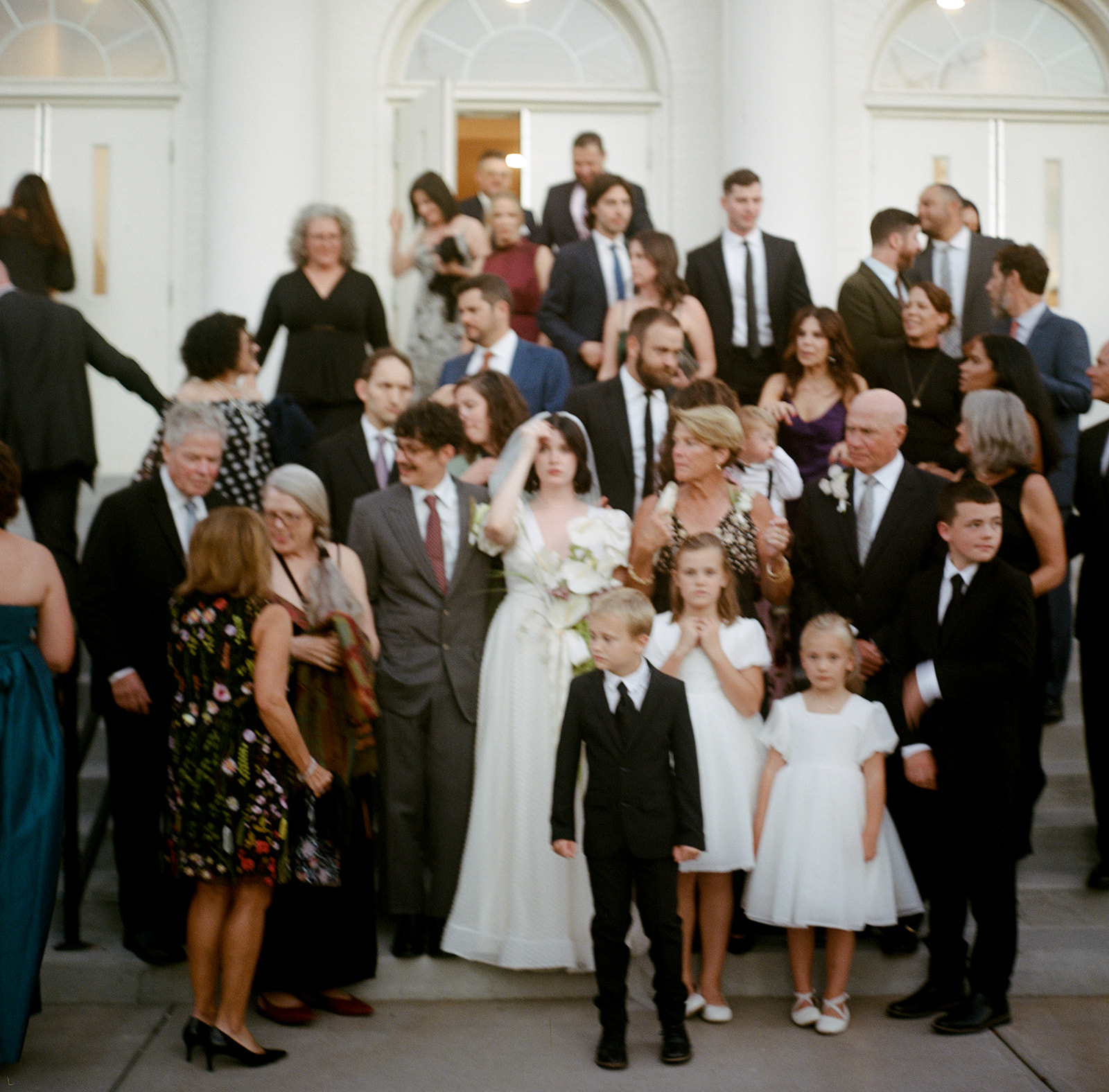 Group of people, including a bride in a white dress, gathered outside a building with white columns.