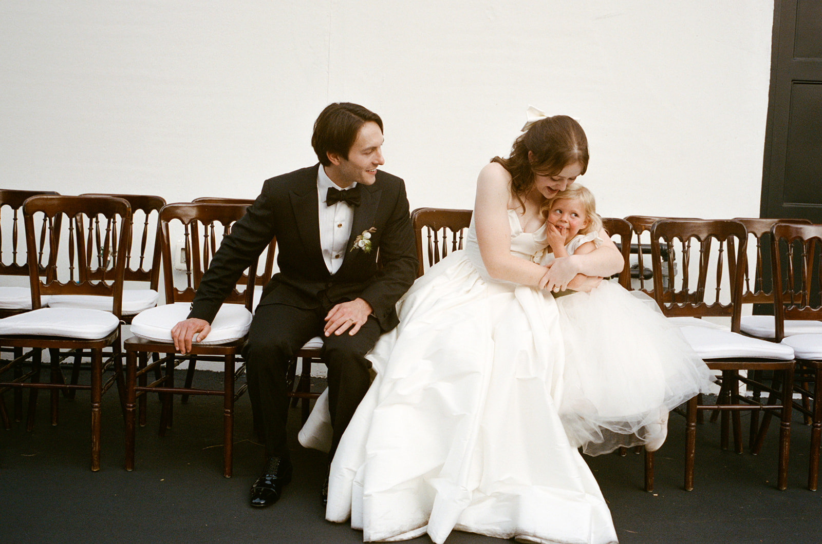 Three people sitting on chairs, two women in wedding dresses and a man in a tuxedo, in a room with chairs.