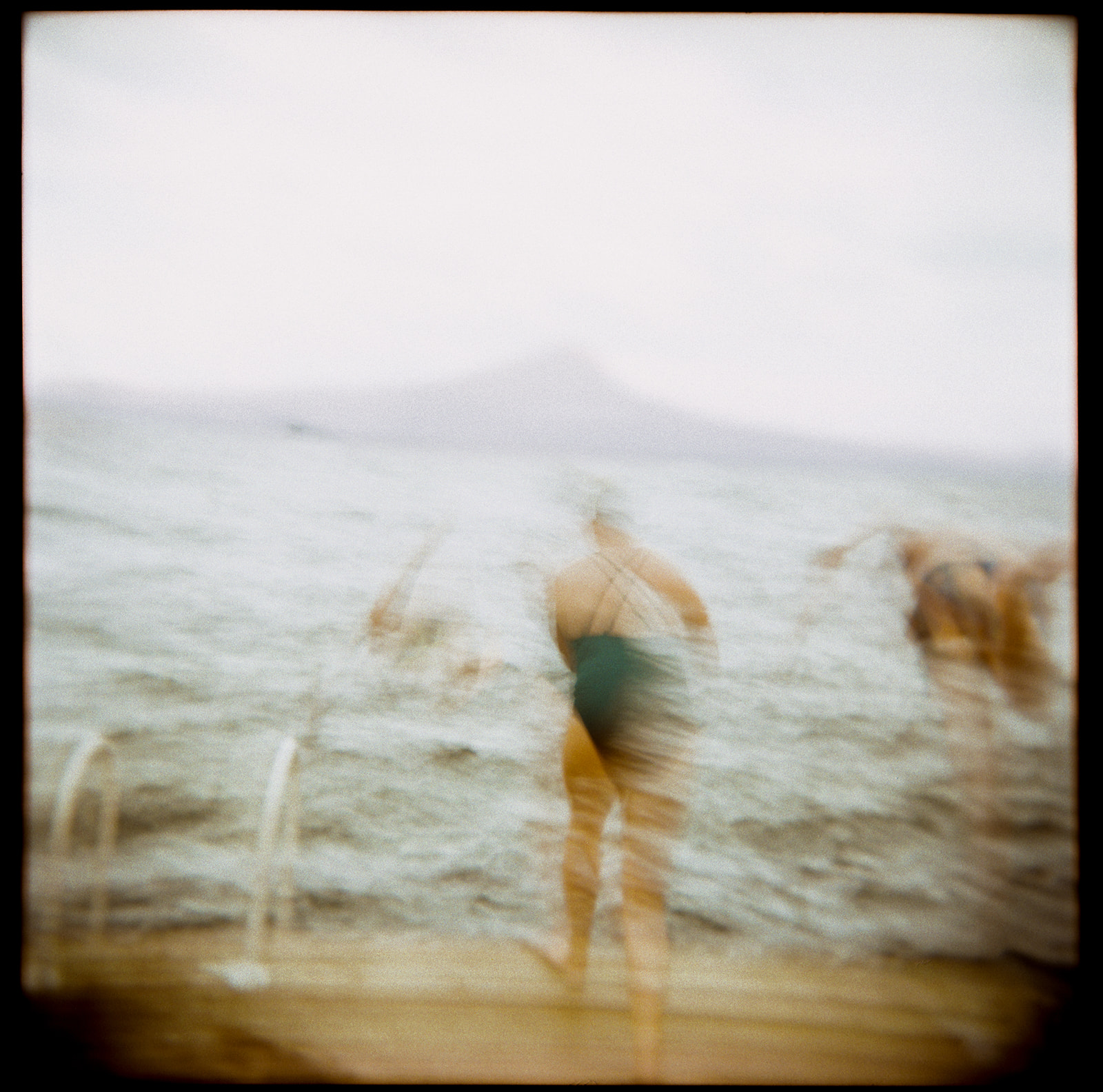 Blurry image of people at the beach, some entering the water, overcast sky, distant land or island in background.