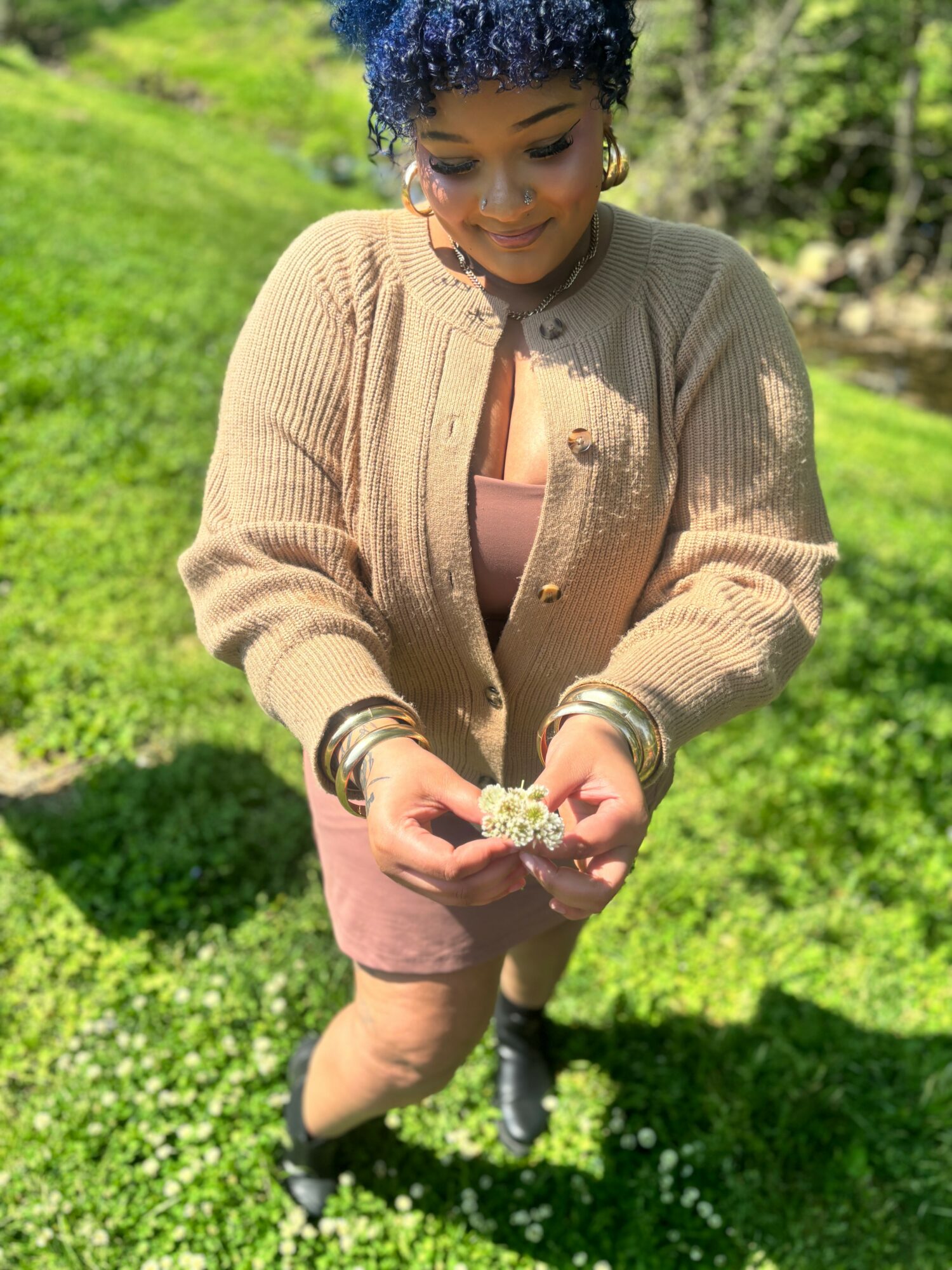 Woman outdoors holding small flower, smiling, wearing beige cardigan and pink dress, surrounded by green grass and trees.