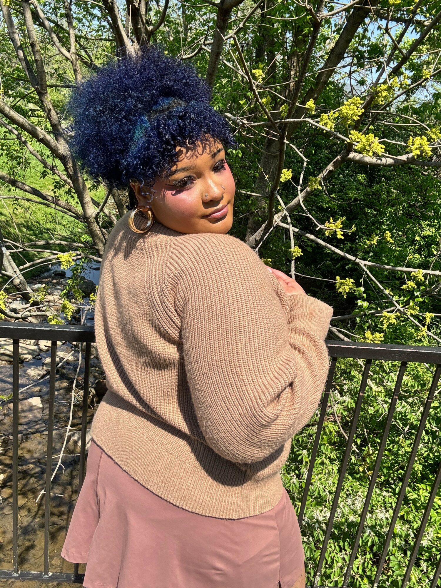 Young woman with curly blue hair stands outdoors near a black metal fence, surrounded by green foliage and yellow flowers.