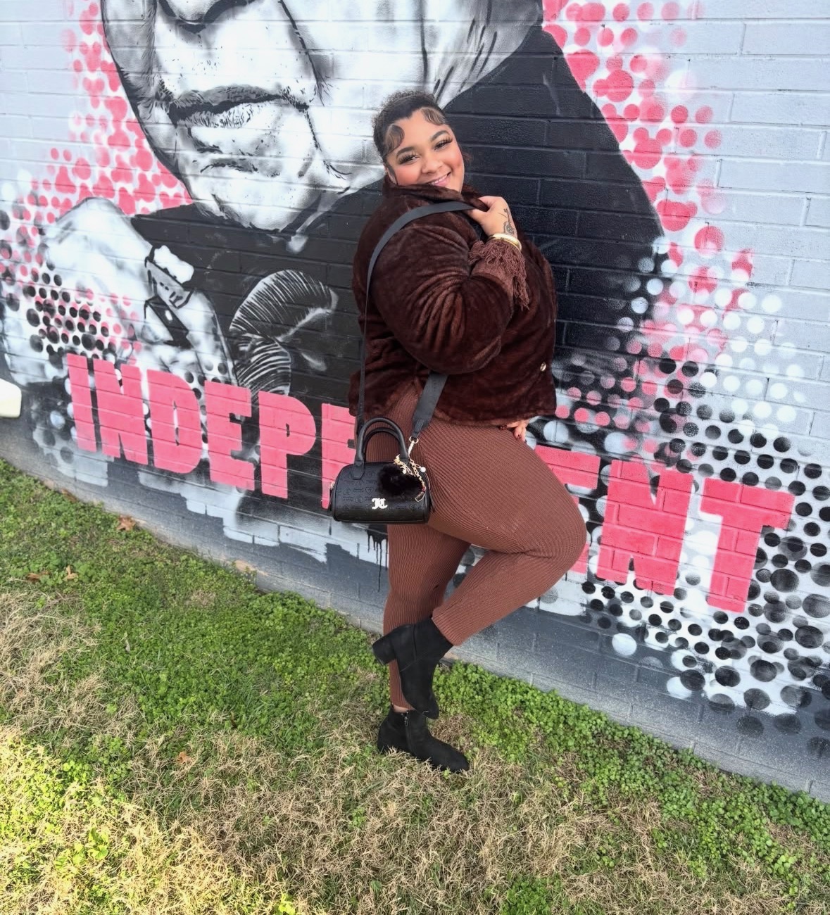 Young woman standing in front of a mural with pink and black dots, smiling, holding a purse, wearing a brown jacket and pants.