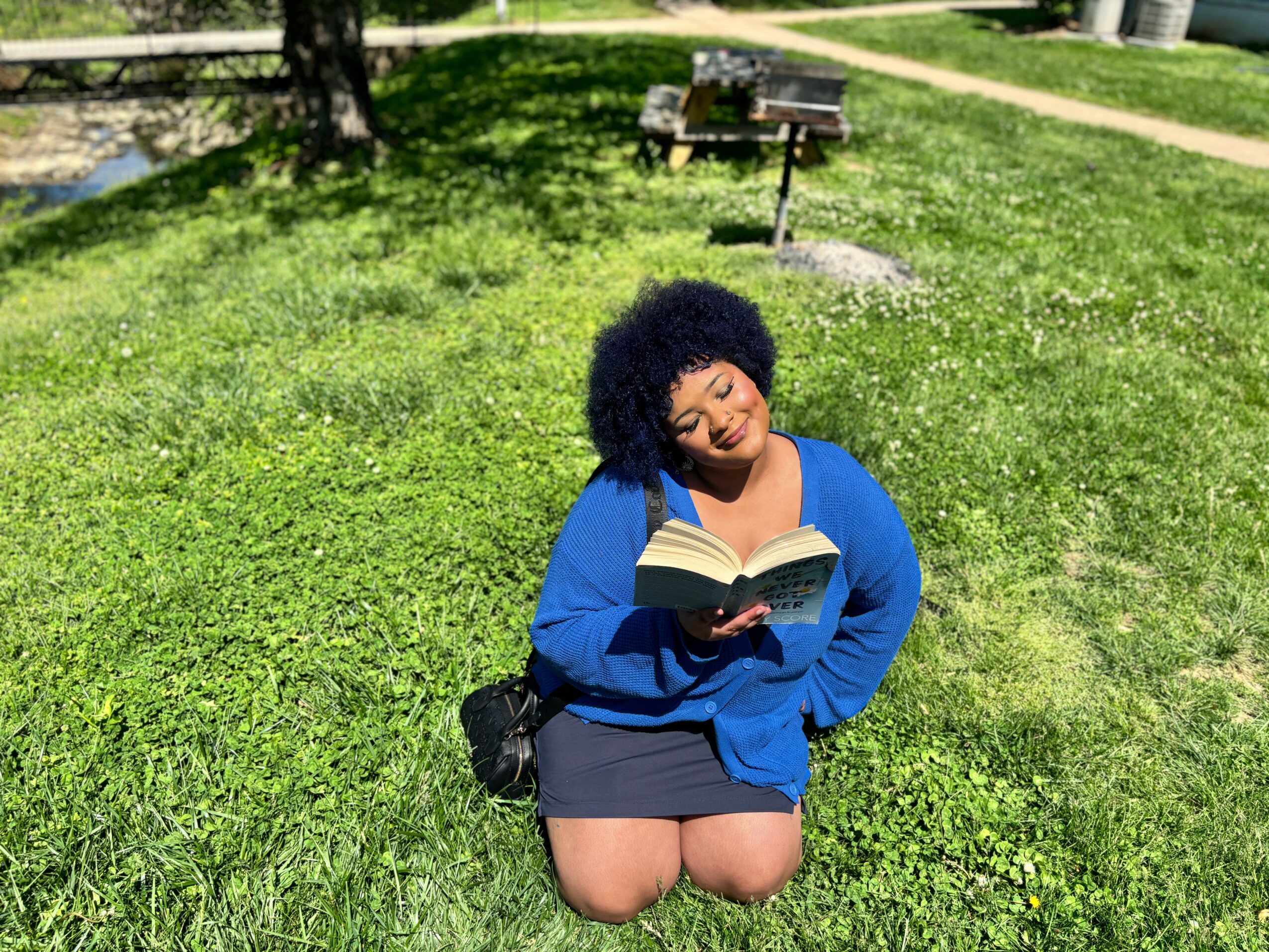 Person with curly hair reading a book outdoors on grass, wearing a blue jacket and black skirt.