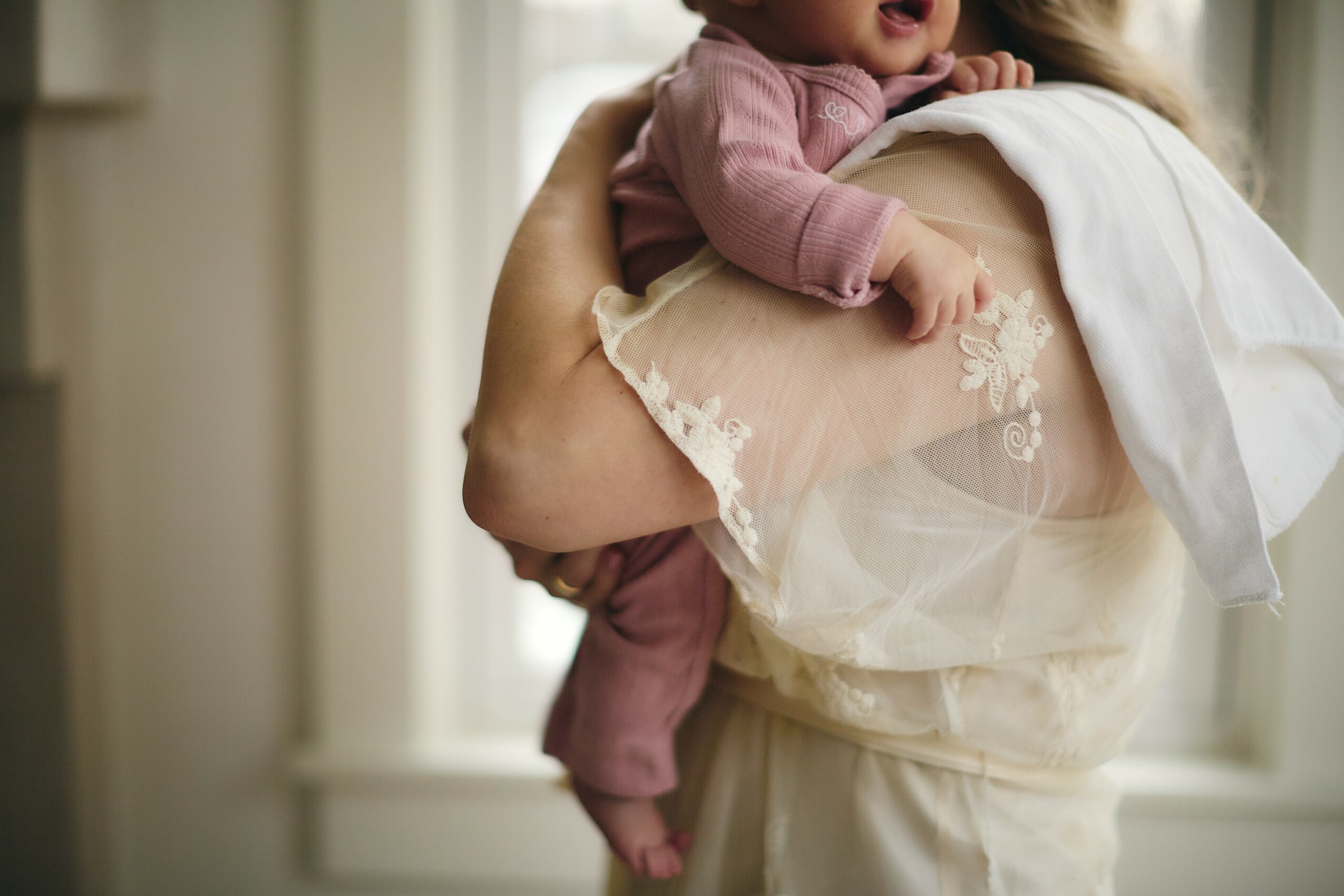Person holding a baby with a window in the background, baby wearing pink clothing, person wearing a white top with embroidery.