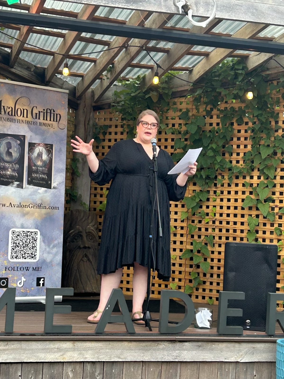 Woman standing on stage, holding papers, speaking with one arm raised, in front of a lattice wall and promotional banner.
