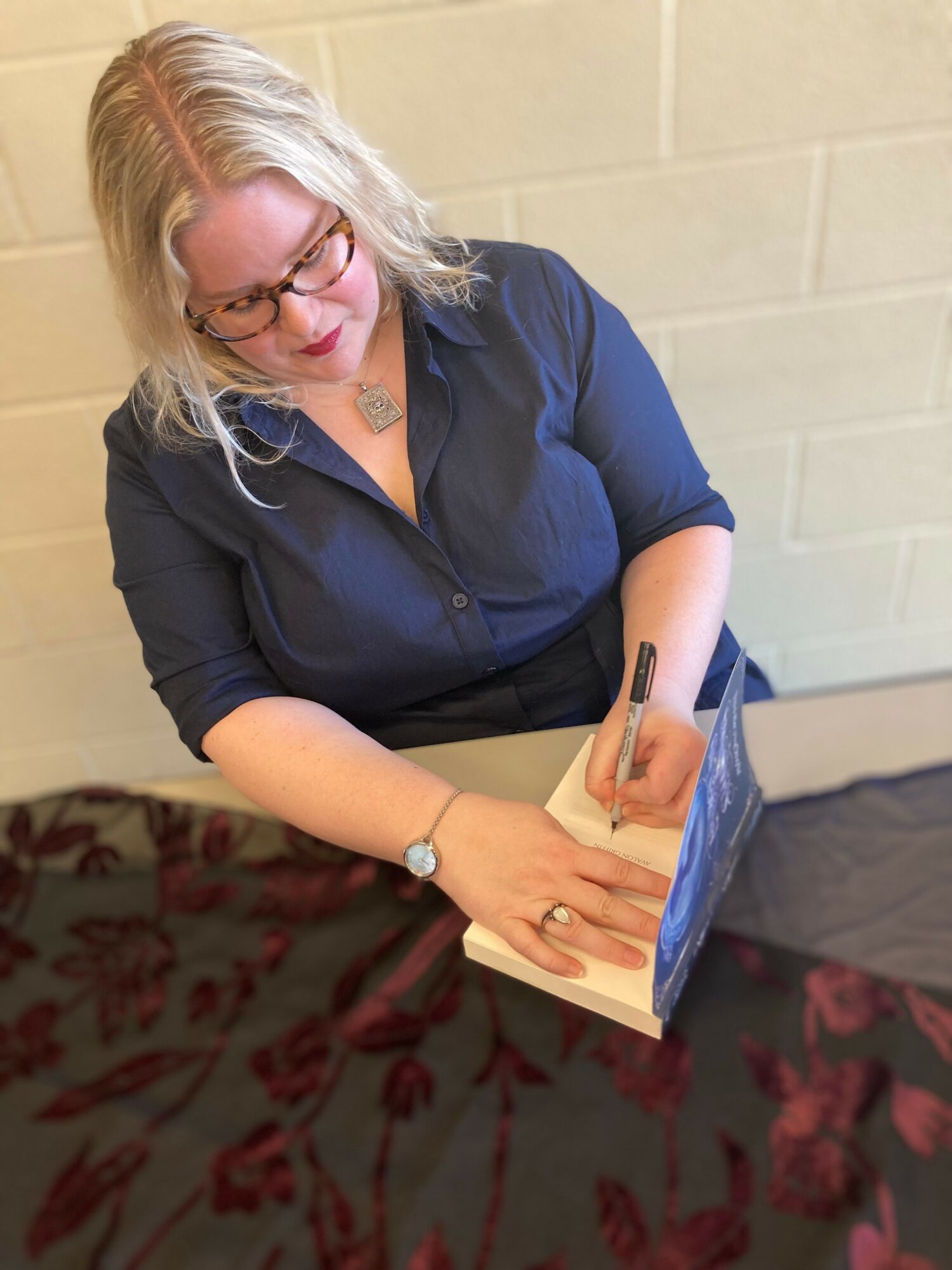 Woman with glasses and blonde hair signing a book at a table with a dark tablecloth.