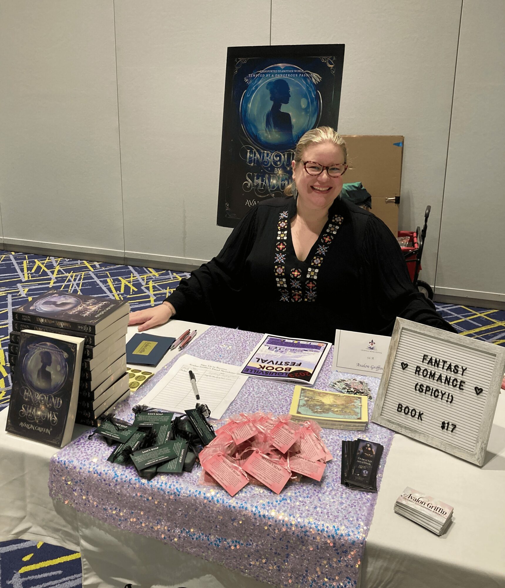 Woman sitting at a table with books and promotional materials, smiling, in a convention setting.