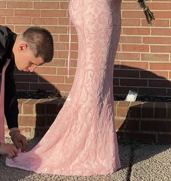 Person kneeling on ground adjusting a pink lace wedding dress outdoors against a brick wall.