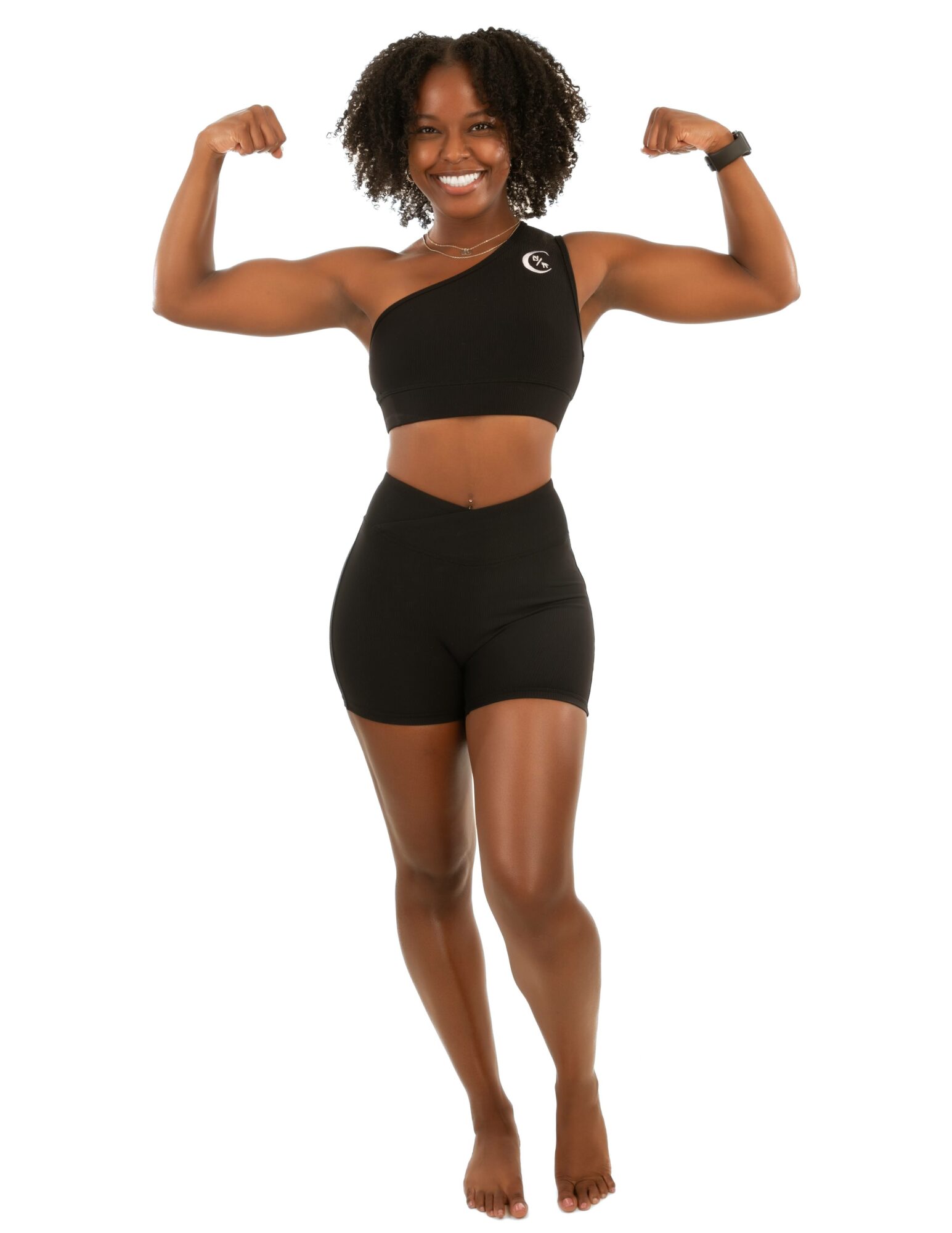 Woman with curly hair flexing arms, smiling, wearing black sports bra and shorts, standing barefoot.