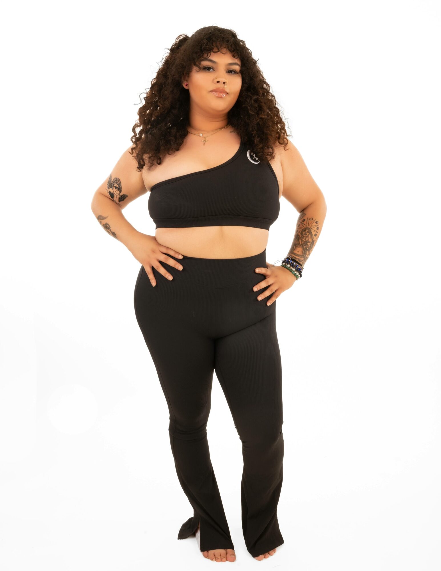 Woman with curly hair wearing a black crop top and high-waisted pants, standing with hands on hips.