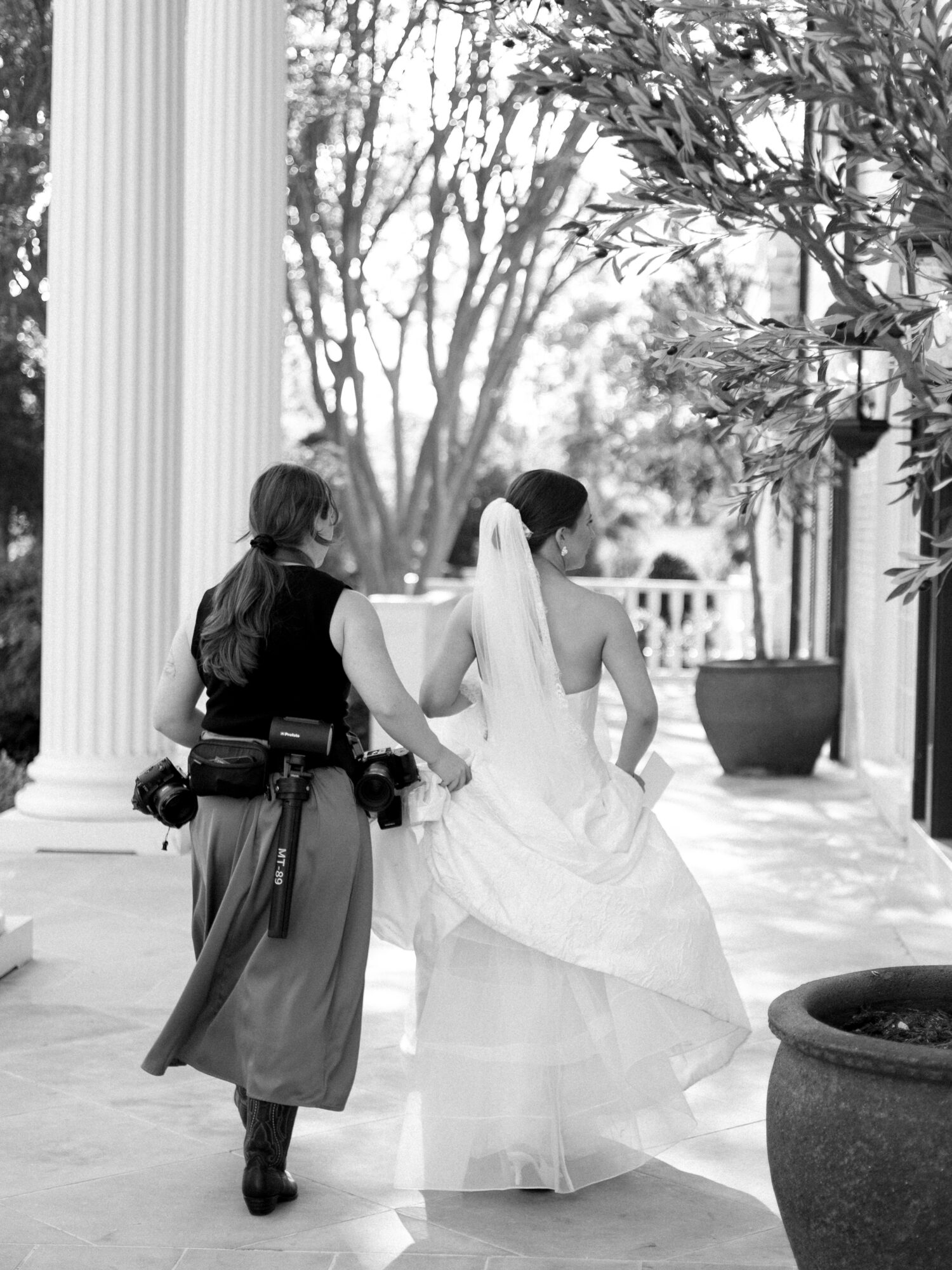 Bride in wedding dress and veil walking with a woman outside near columns and trees.