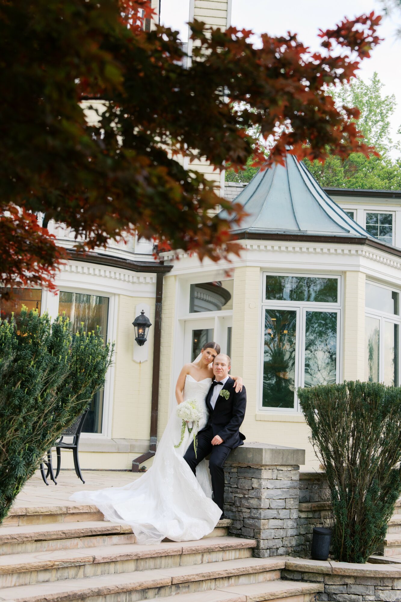 Bride and groom standing on steps outside a building with large windows and a turret, surrounded by bushes and trees.
