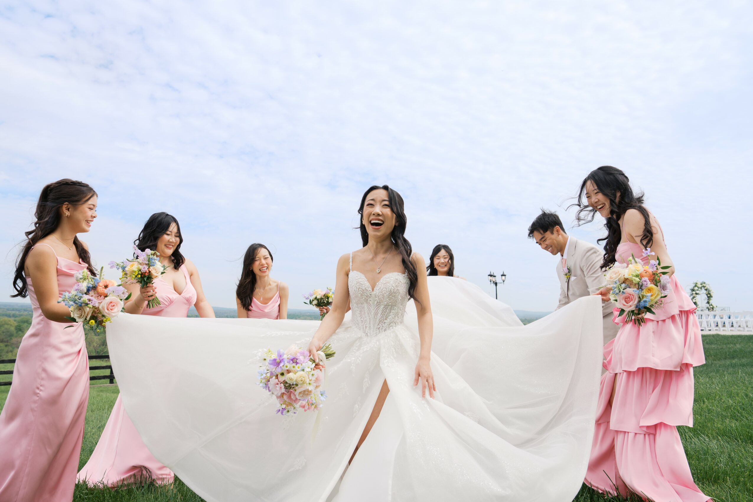 Bride in white dress holding bouquet, surrounded by bridesmaids in pink dresses on green field under cloudy sky.