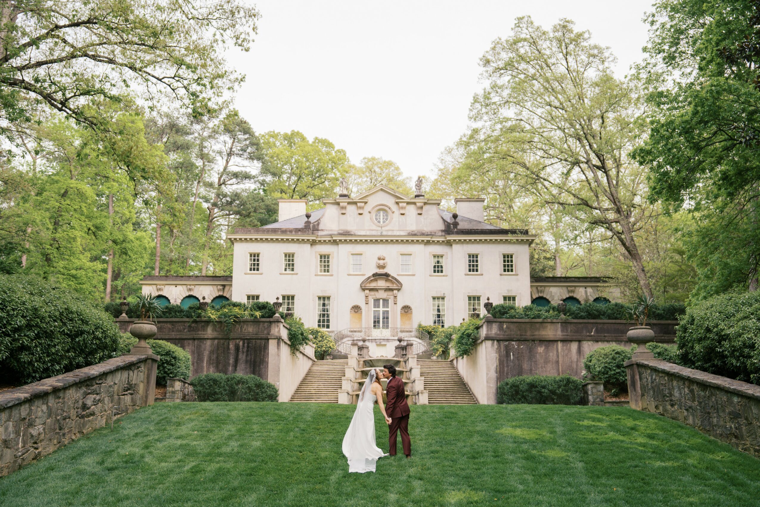 A couple stands on a lawn in front of a large white mansion surrounded by trees.