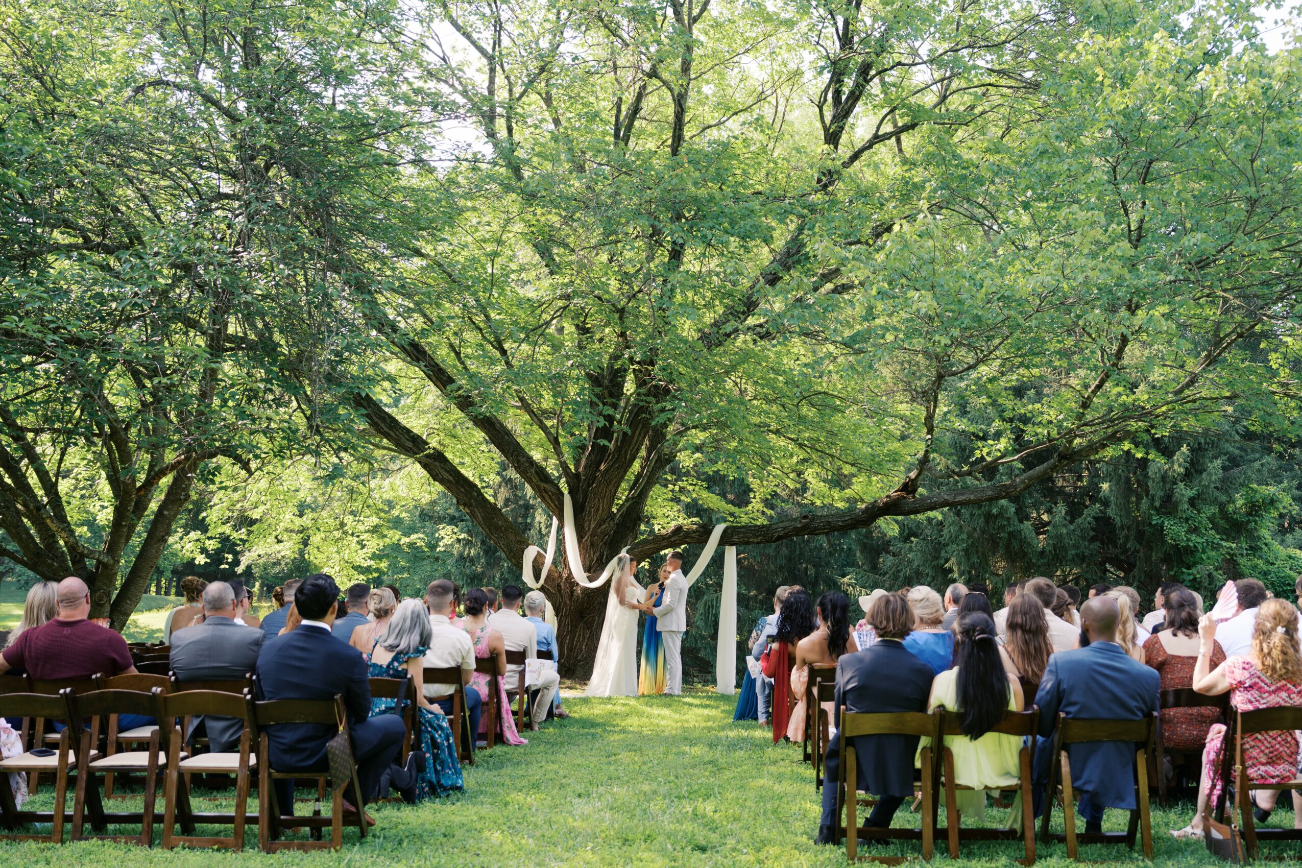 Outdoor wedding ceremony with guests seated under trees, bride and groom standing at altar, lush green foliage.