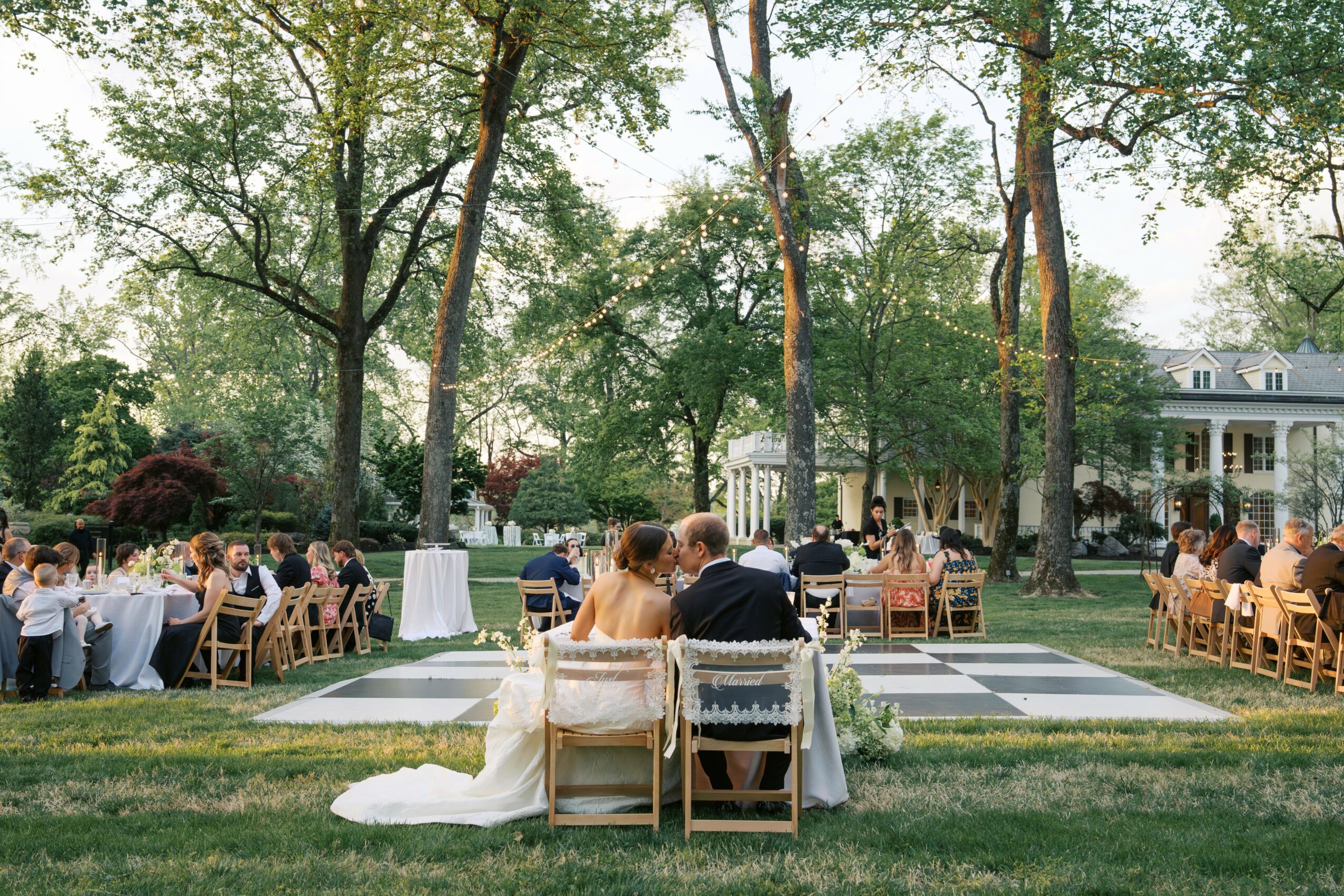 Outdoor wedding reception with tables, chairs, and a dance floor surrounded by trees and a building in the background.