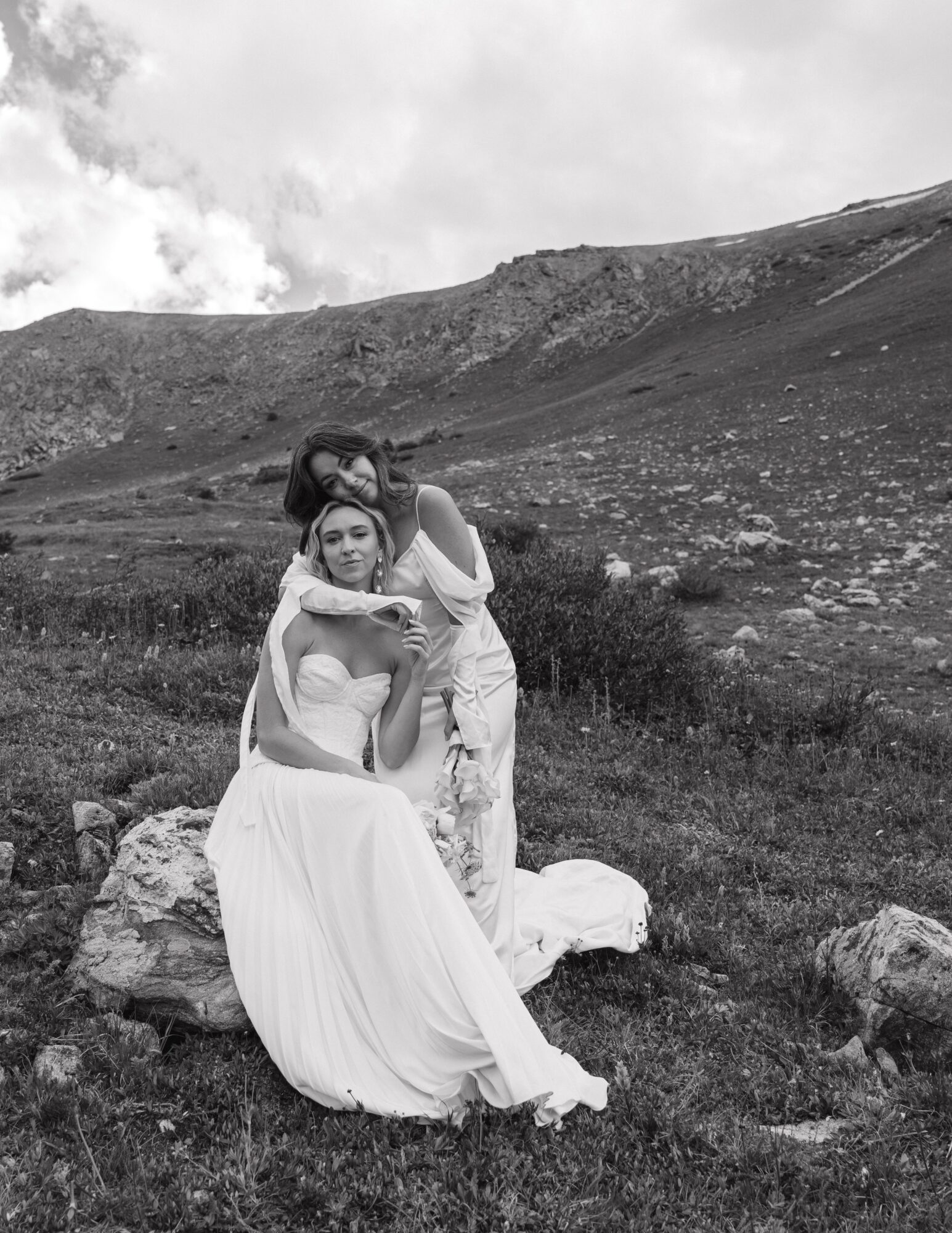 Two women in white dresses sitting and standing outdoors on rocky terrain with a mountain and cloudy sky in background.