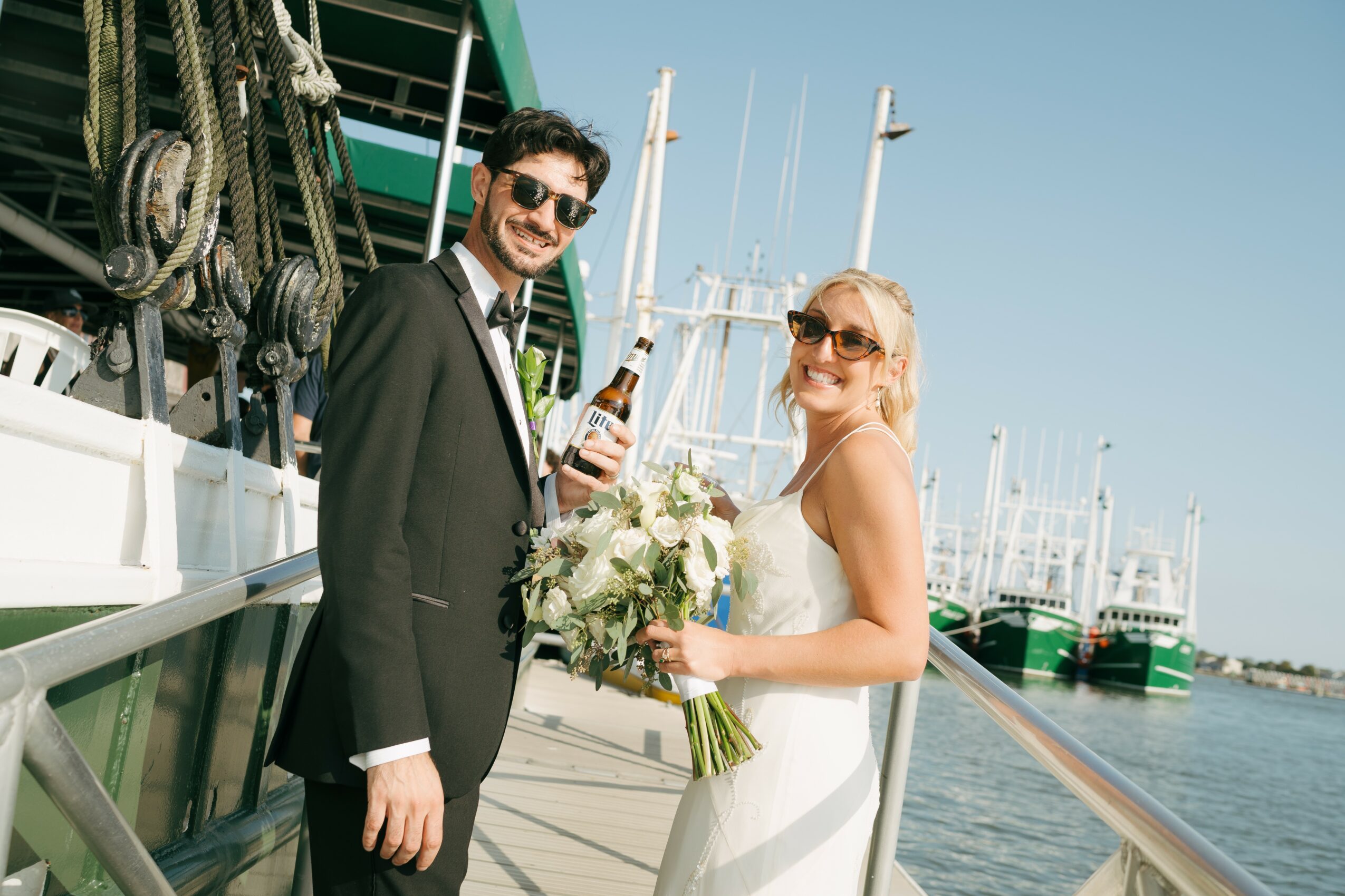 A smiling couple in wedding attire stands on a dock near boats, holding a bouquet and a bottle of champagne.