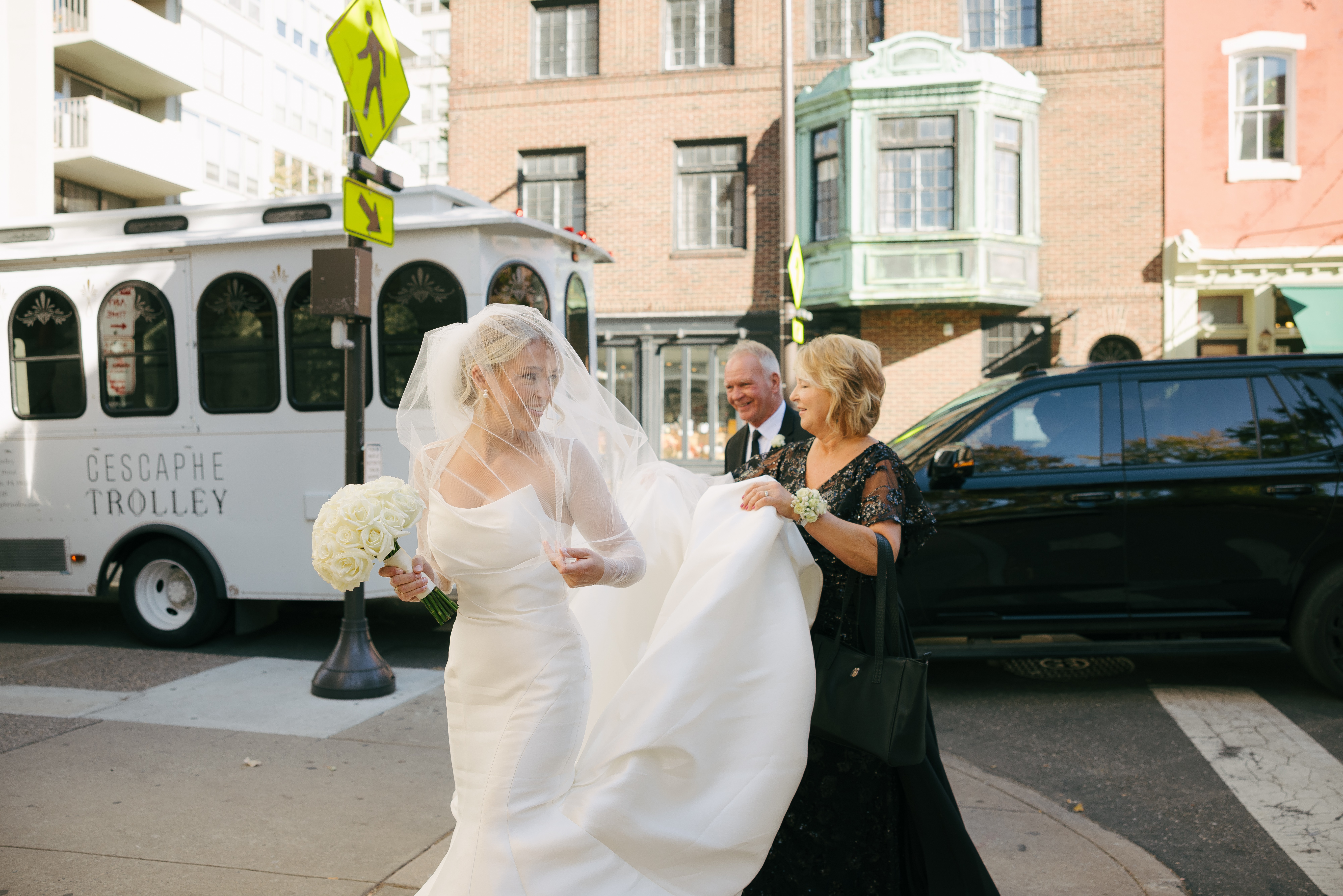 Bride in white dress holding bouquet, woman in black dress holding train, two men in suits, city street background with trolley and parked cars.