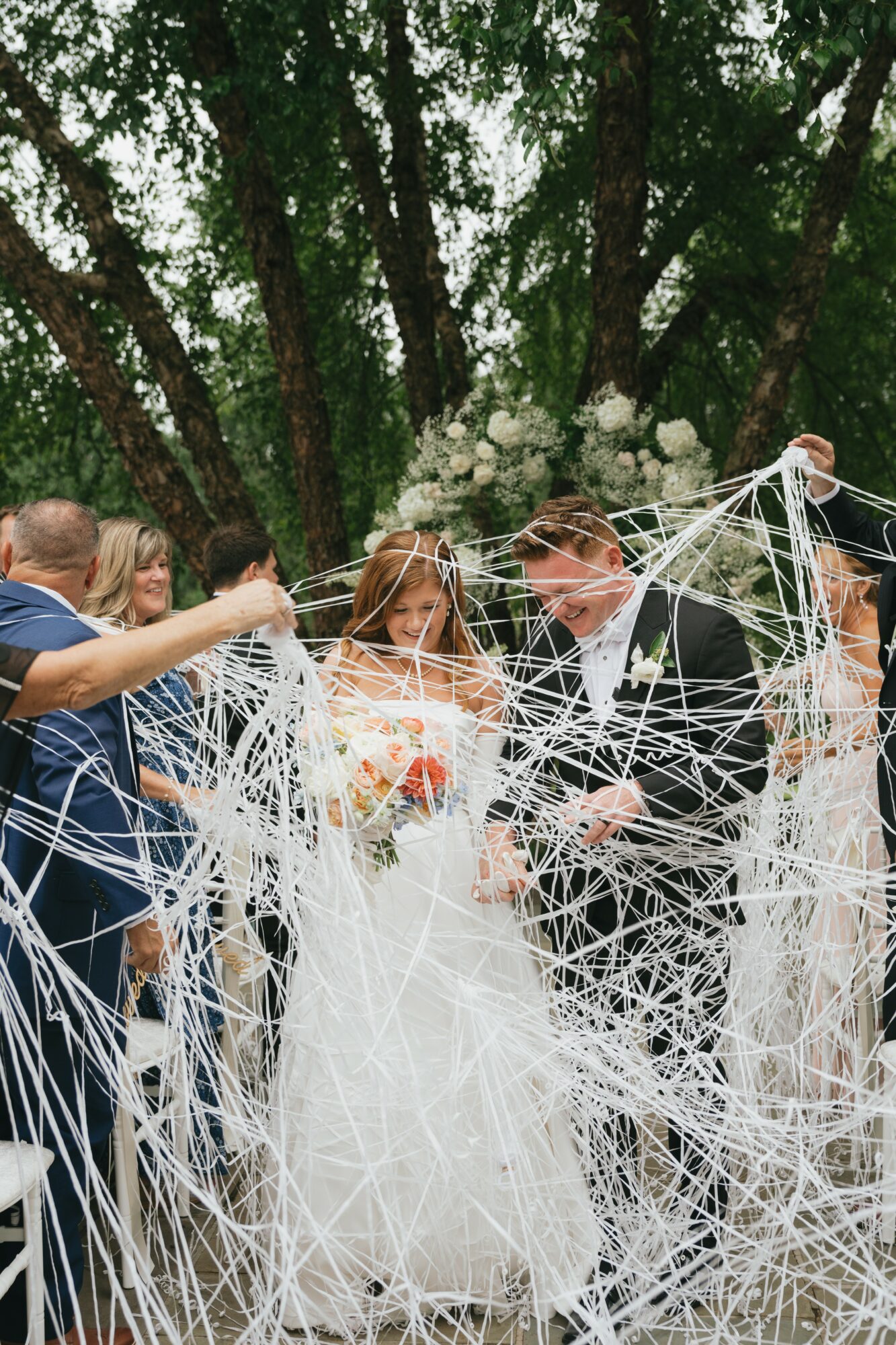 Bride and groom holding hands, surrounded by friends, with trees and white flowers in background, during wedding celebration.