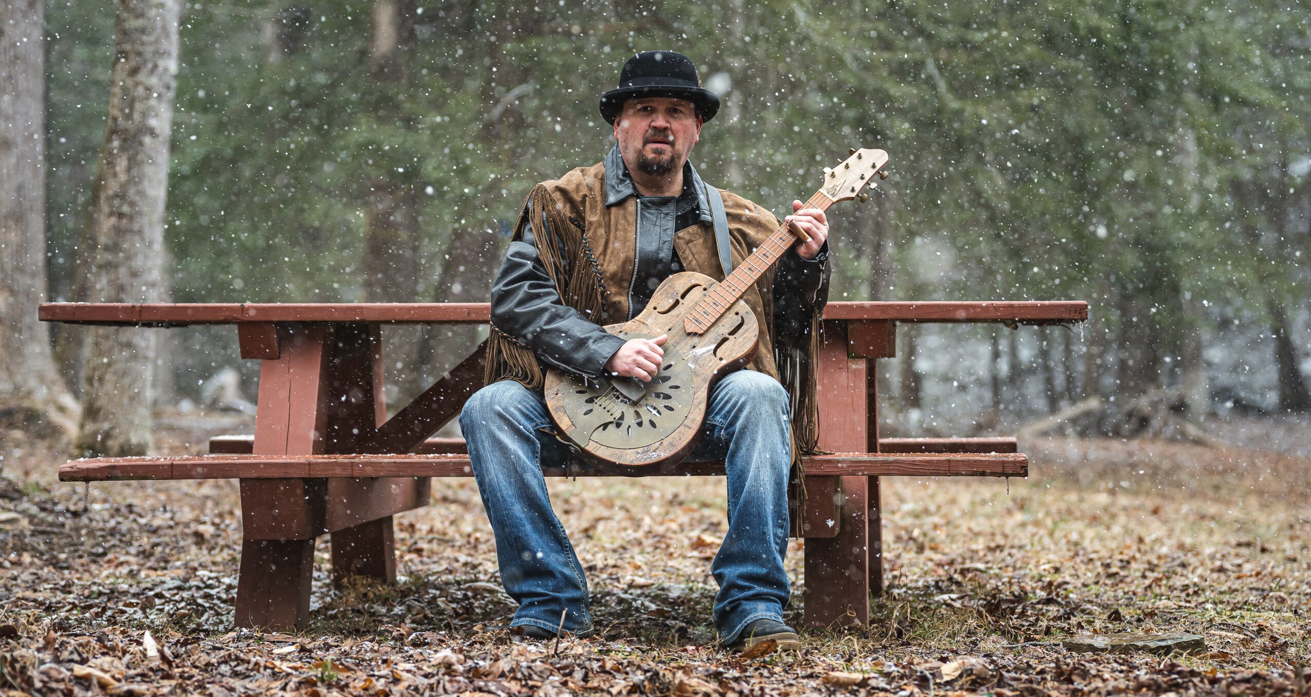 Man sitting on a bench outdoors playing a guitar, surrounded by trees and fallen leaves.
