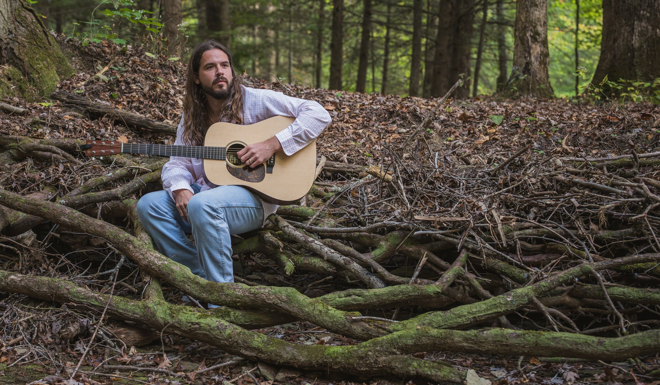 Man with long hair and beard sitting on fallen logs in a forest, playing an acoustic guitar.