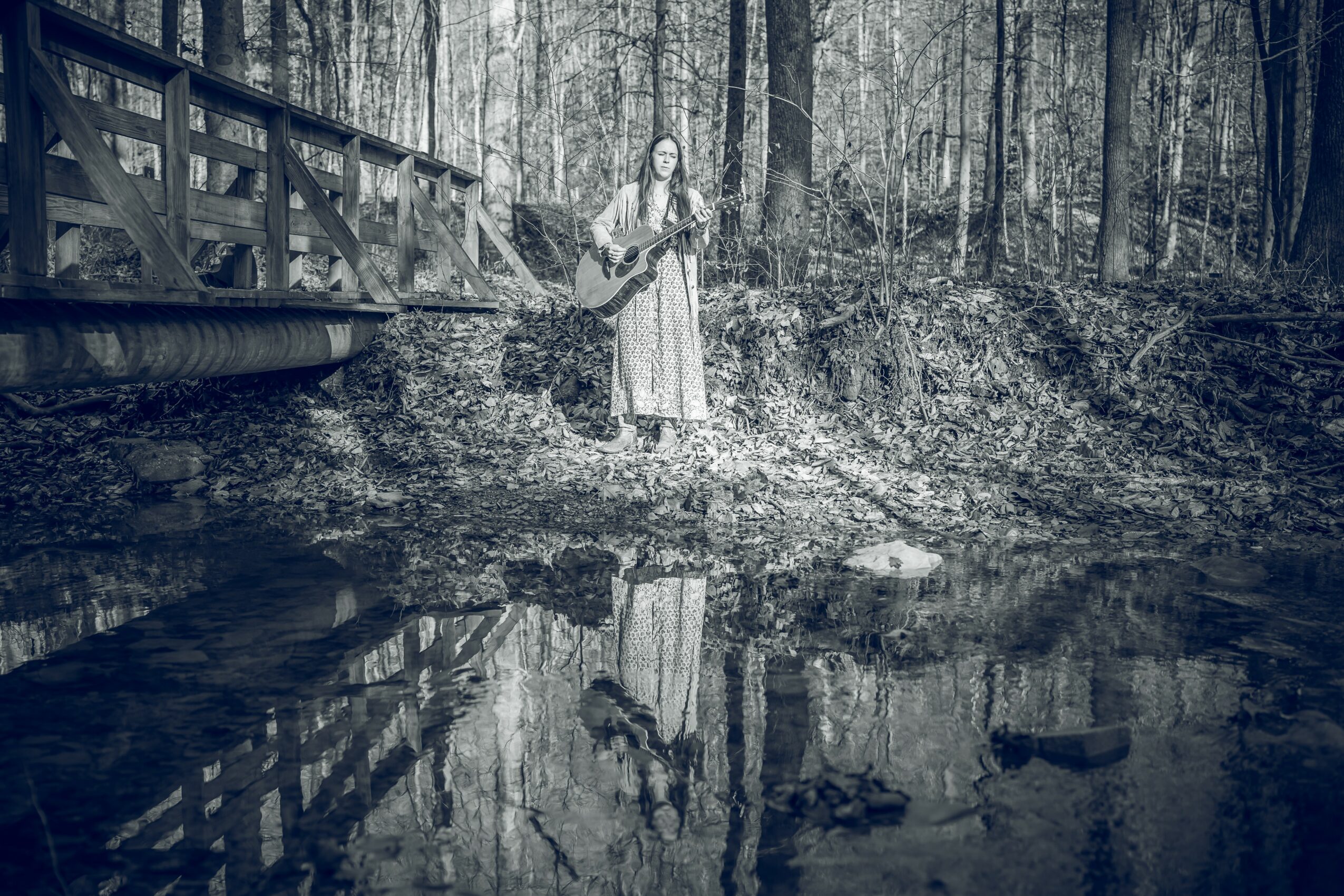 Person standing on a wooded riverbank holding a guitar, with a bridge nearby and trees in the background.