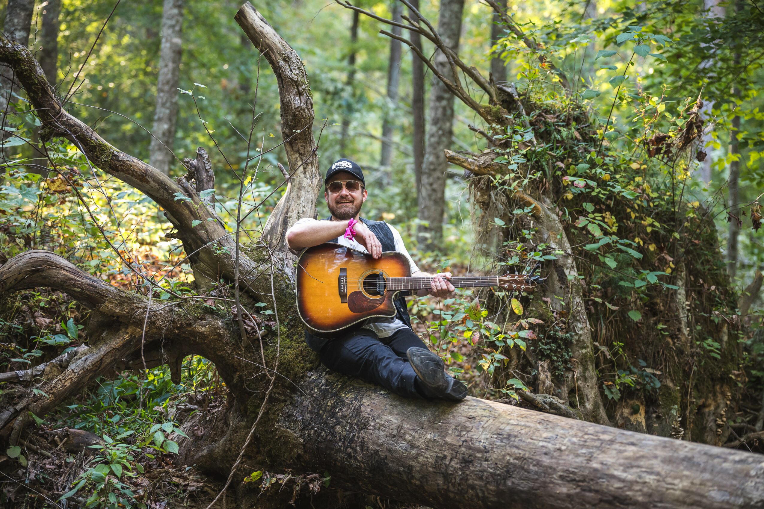Person sitting on a fallen tree in a forest, playing an acoustic guitar, surrounded by green foliage.