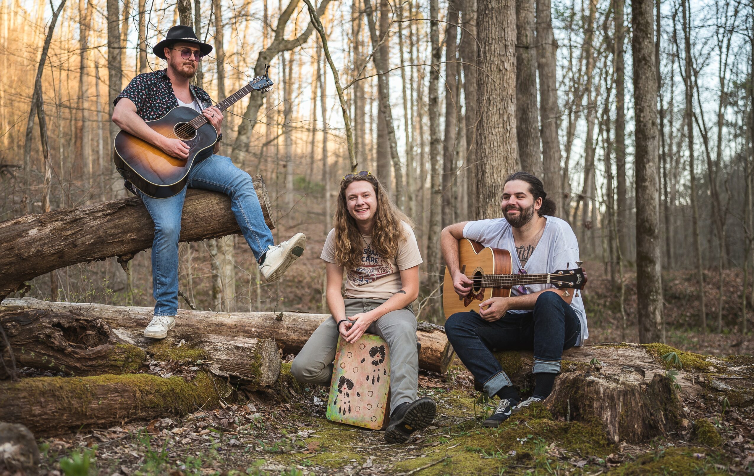 Three people in a forest, two playing guitars and one sitting on a drum, smiling and enjoying music.