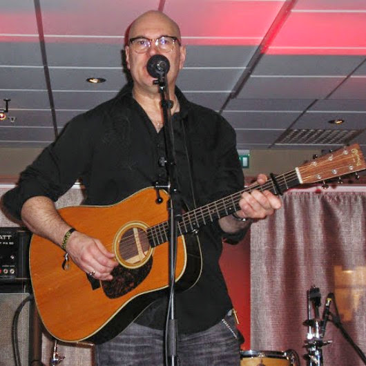 Man with glasses playing acoustic guitar and singing into a microphone indoors.