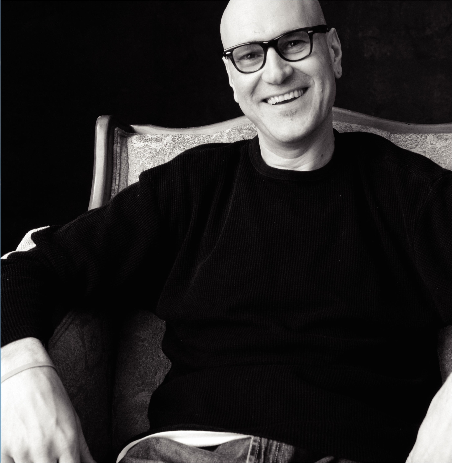 Man with glasses smiling, sitting on a chair, wearing a black sweater, in black and white.